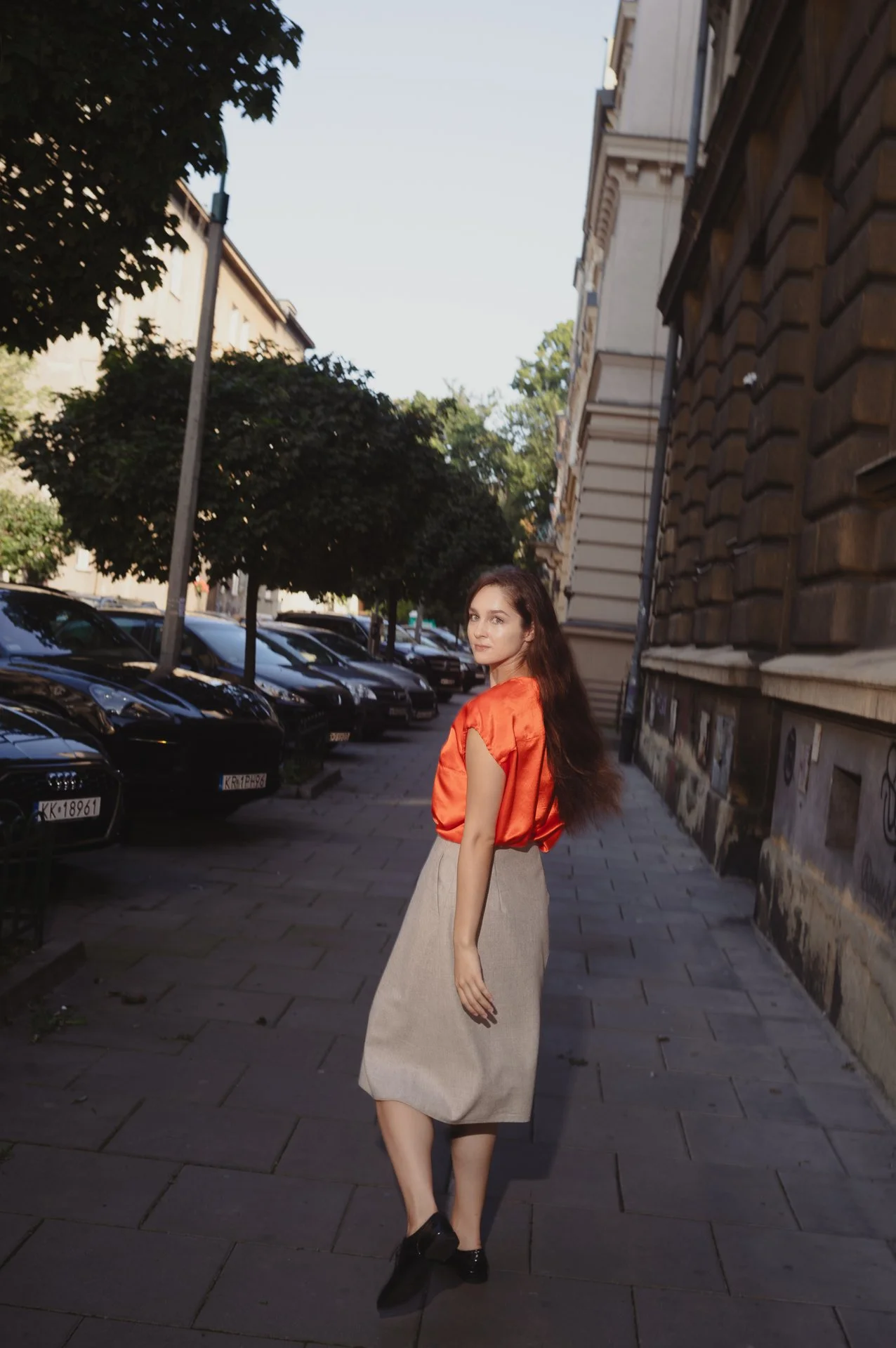 Woman in red blouse and beige skirt walking on city sidewalk with parked cars and buildings.
