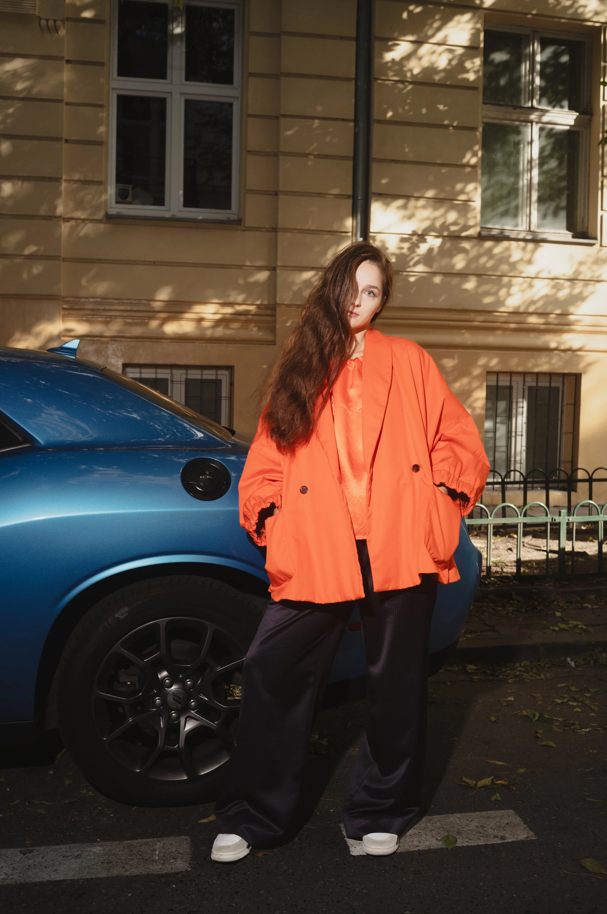 Woman in an oversized orange coat and dark pants standing next to a blue car on a street with a beige building in the background.