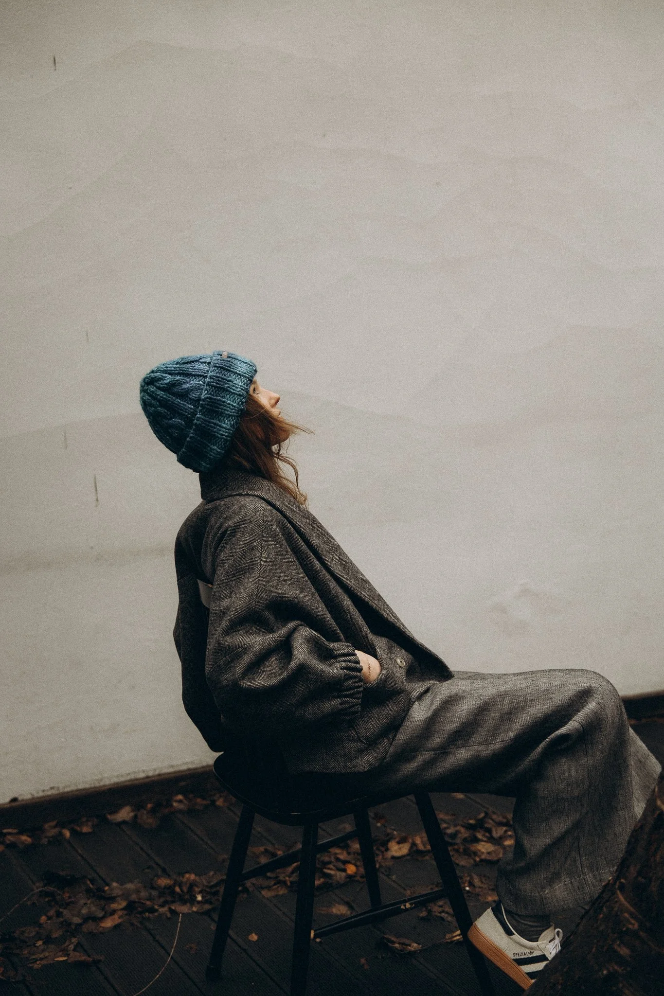 Person in gray outfit and blue knit hat sitting on a chair against a white wall, surrounded by dried leaves on the ground.