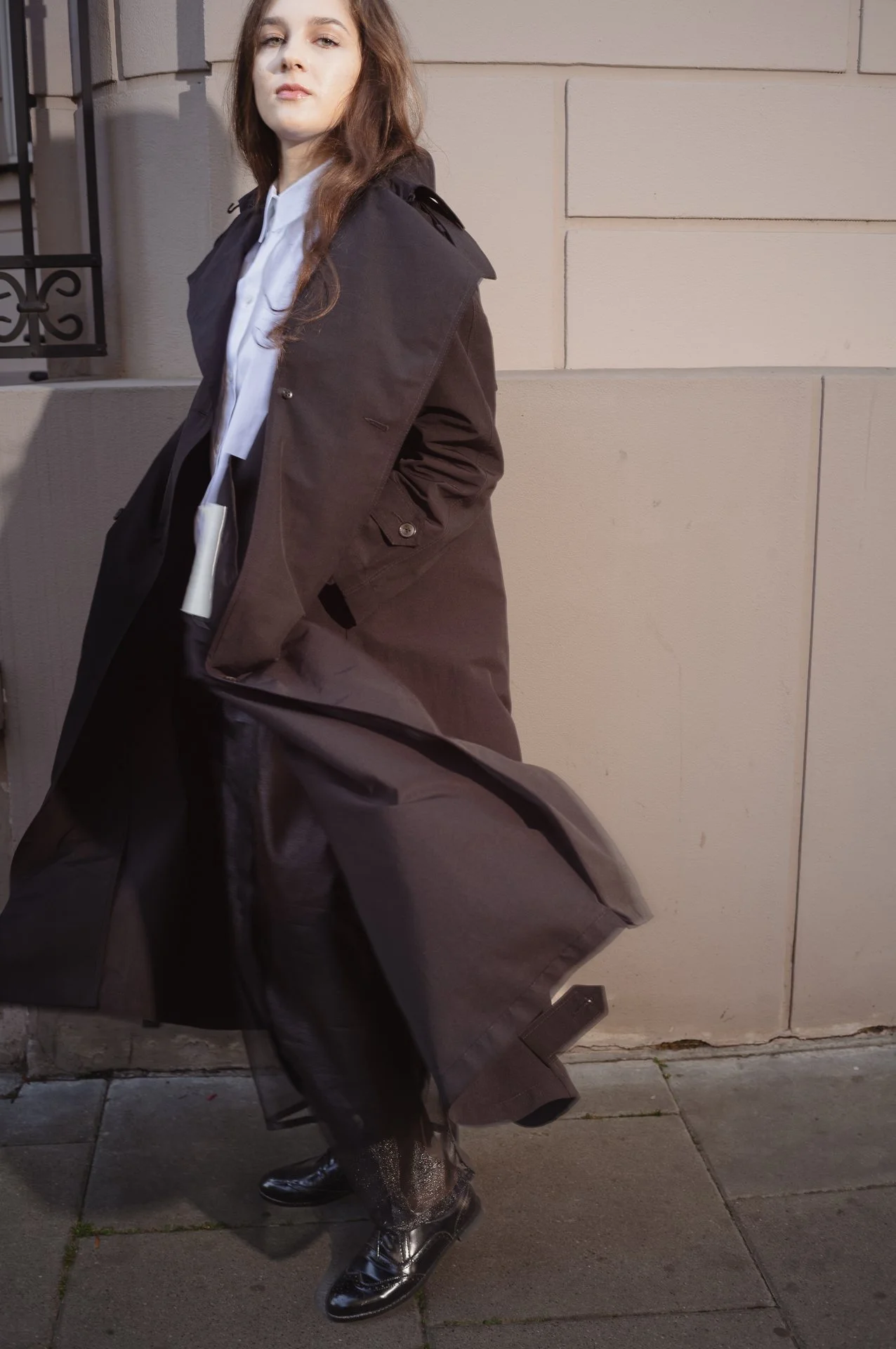 Person wearing a long black coat and patent leather shoes outside a building.