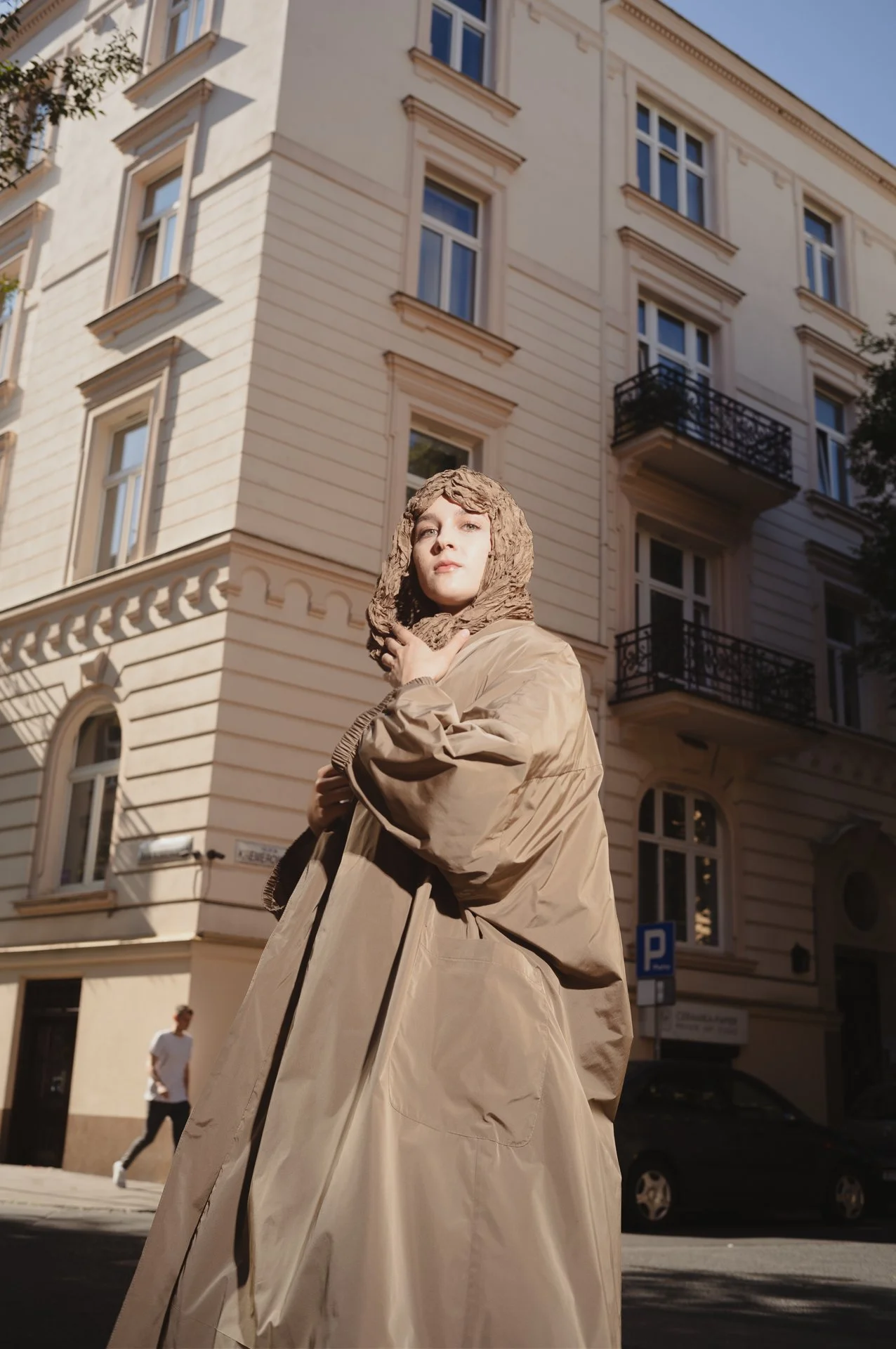 Person in a tan oversized coat standing in front of a European-style building during the day.