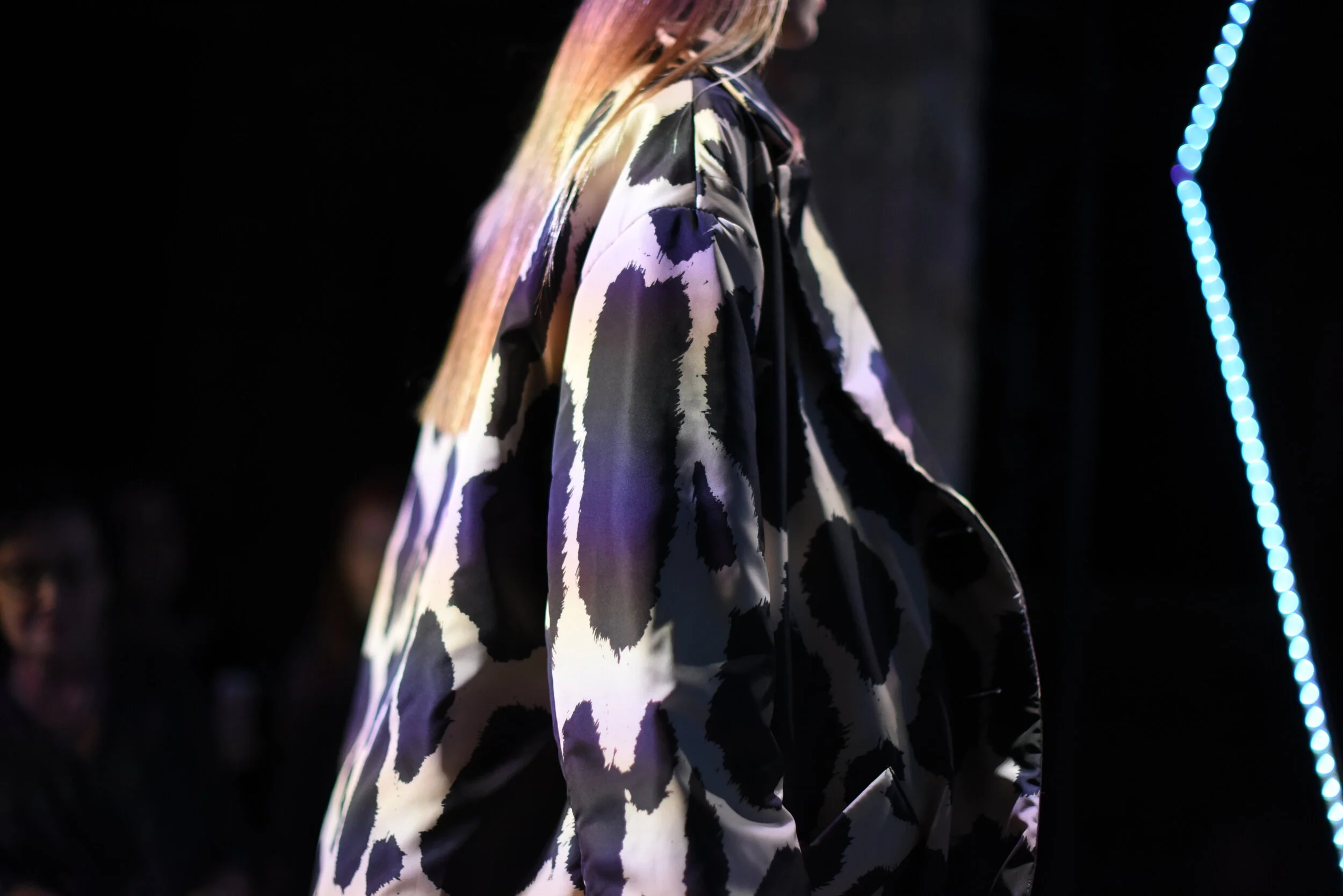 Person wearing a loose, abstract-patterned coat on a fashion runway with a dark background and blue vertical lights.