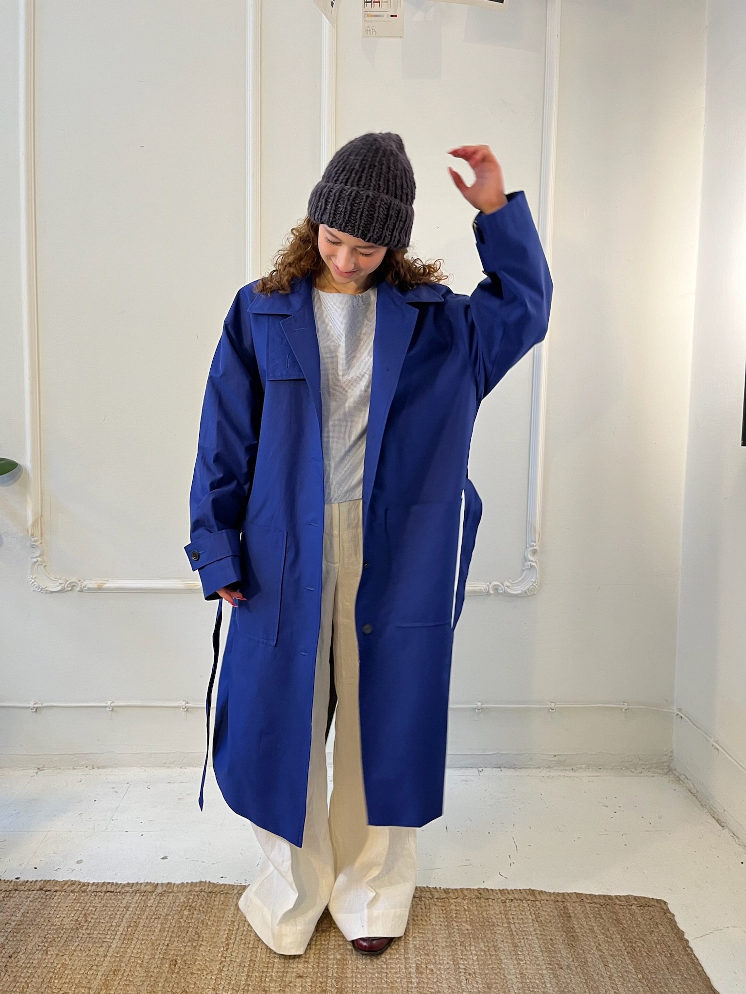 Person wearing a blue trench coat over a white outfit and a gray knit beanie in a minimalist room.