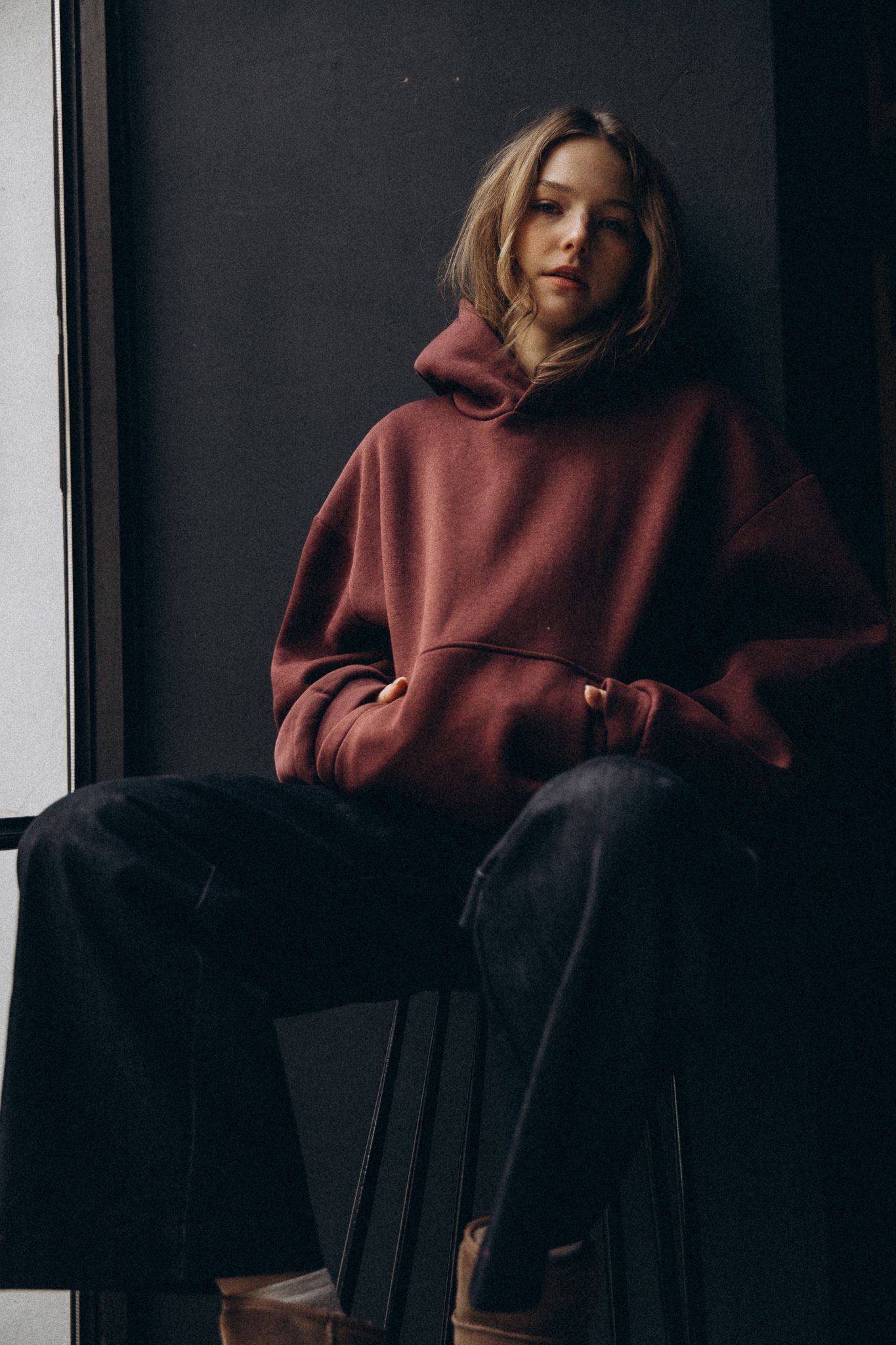 Person wearing a maroon hoodie and black pants, sitting casually on a stool against a dark background.