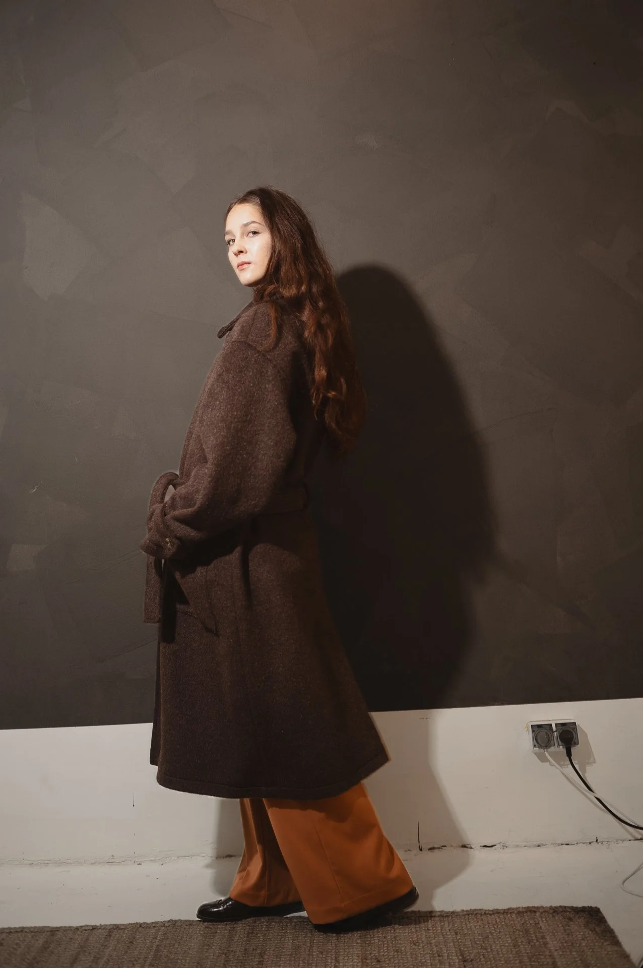 Person in a long brown coat and orange pants standing indoors against a dark wall.