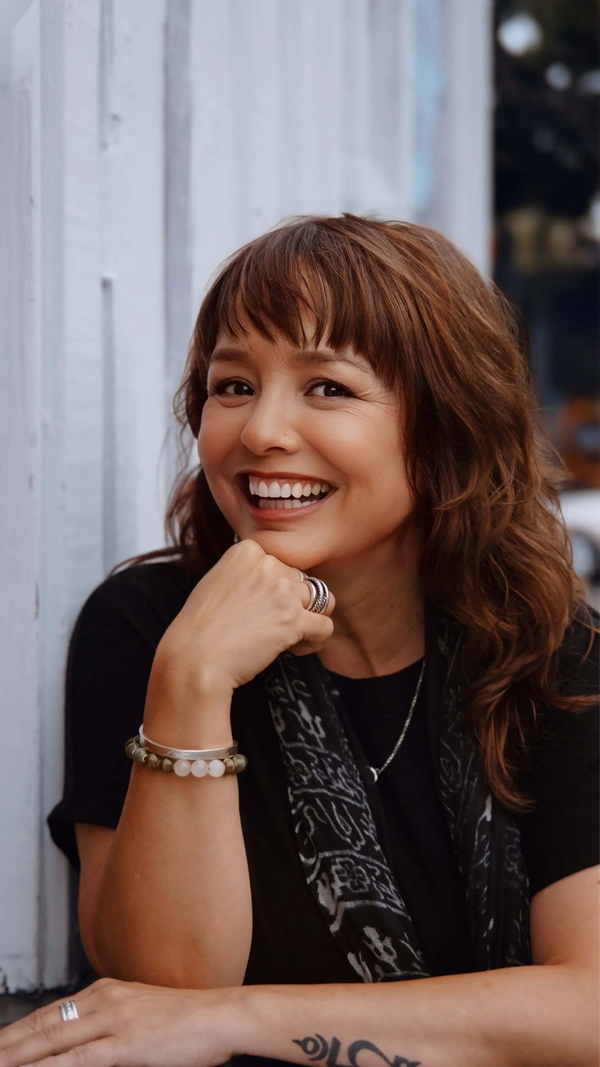 A woman with brown hair and a big smile, sitting against a light-colored wooden wall, wearing rings, bracelets, and a black top.