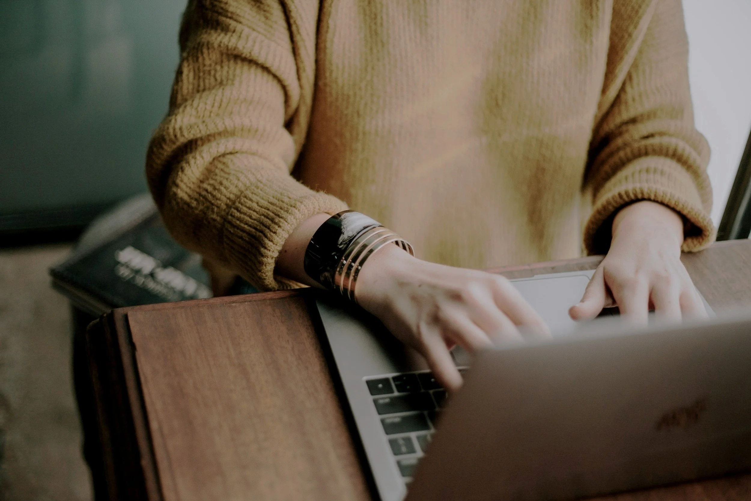 Person wearing a brown sweater typing on a laptop with a wooden surface nearby.