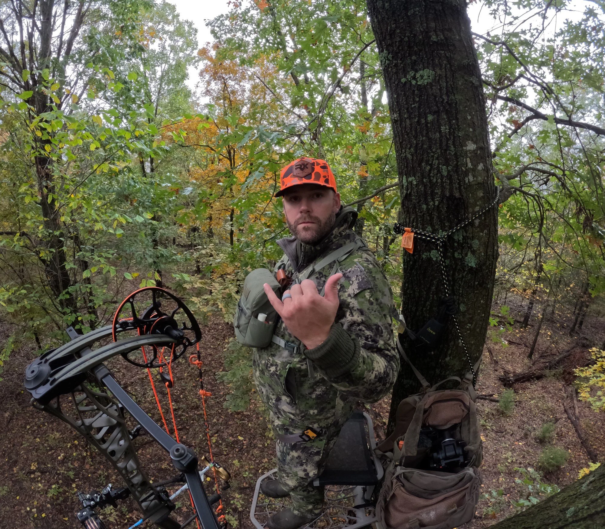 Hunter in orange cap in a tree stand with his bow, giving a hang loose sign
