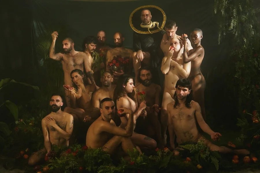 Group of naked people posing among greenery, with one person holding a framed picture of a man's face with a snake around his neck above them. The scene appears staged with dramatic lighting.