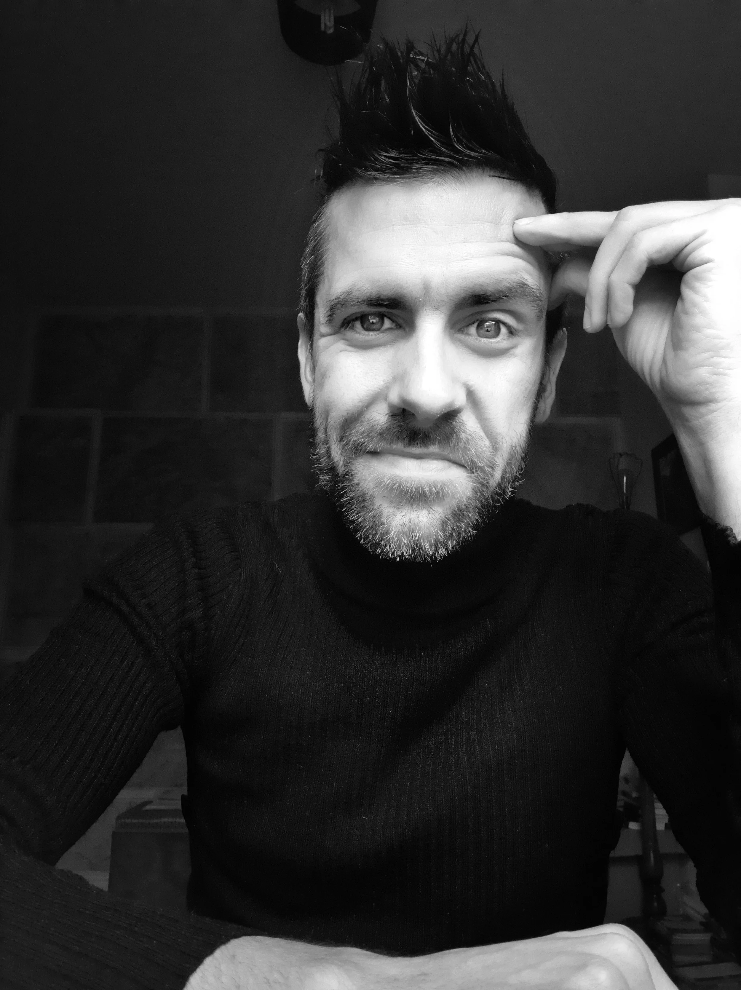 A black and white portrait of a man with short spiky hair and a beard, looking directly at the camera, resting his head on his hand.