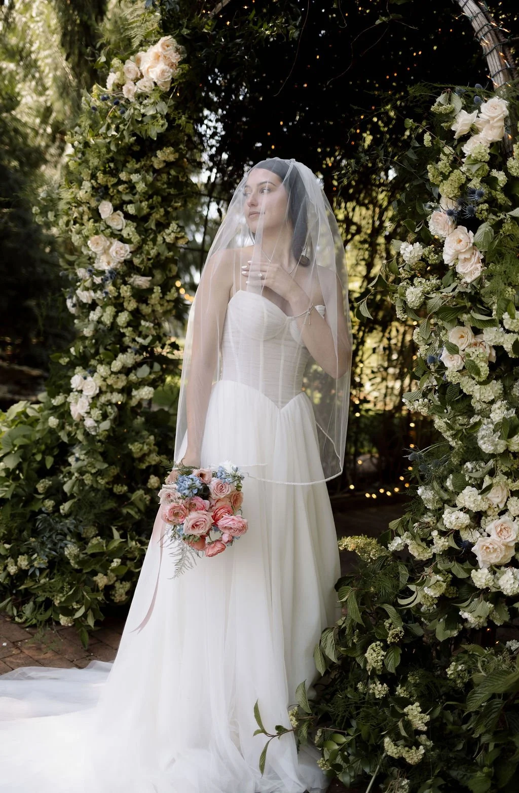 Bride standing next to outdoor wedding floral arch designed with spring viburnums and roses.