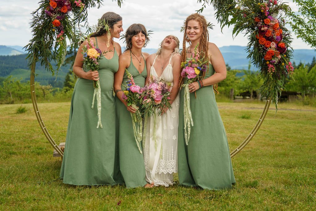 Four women standing outdoors on a grassy field, smiling and laughing under a large floral ring arch. The women are dressed in long green dresses, except for one in a white dress. They hold bouquets of flowers. The background features green hills and trees under a partly cloudy sky.
