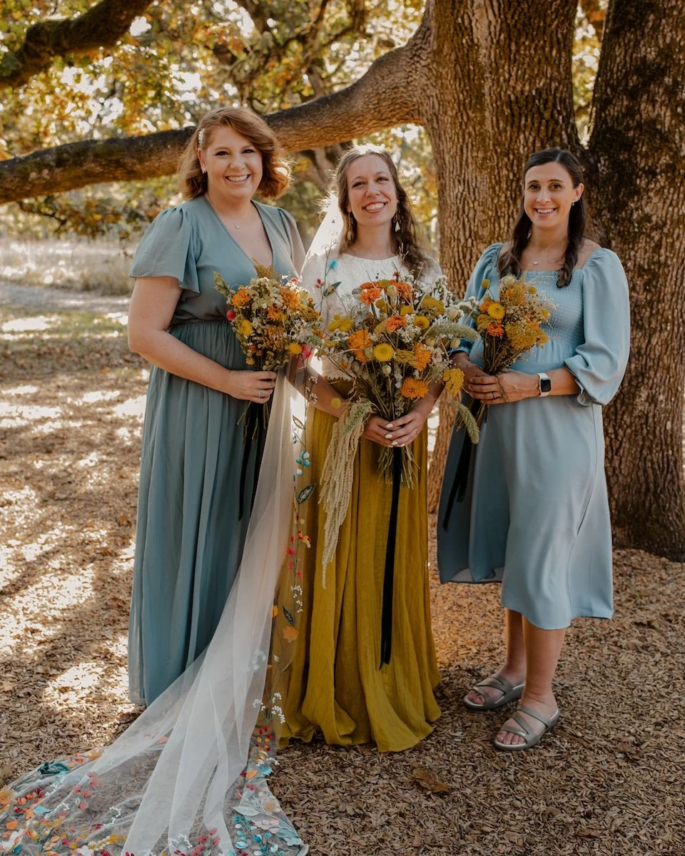 Three women standing outdoors near a large tree during autumn, holding bouquets of yellow and orange flowers, smiling for a photo.