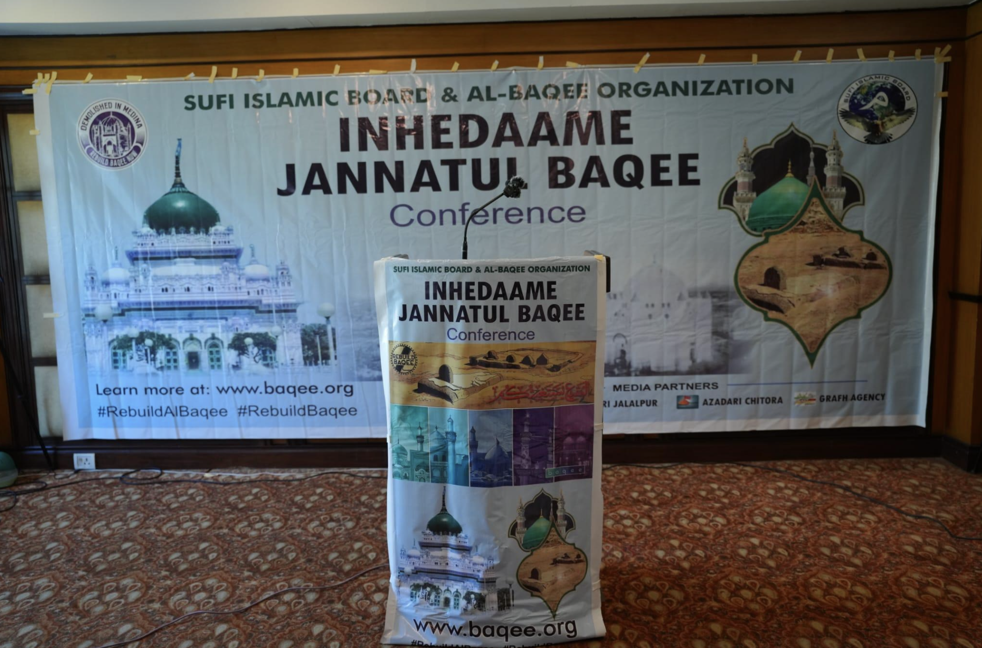 An indoor press conference with a purple banner in the backdrop reading "INHEDAAME
JANNATUL BAQEE CONFERENCE"