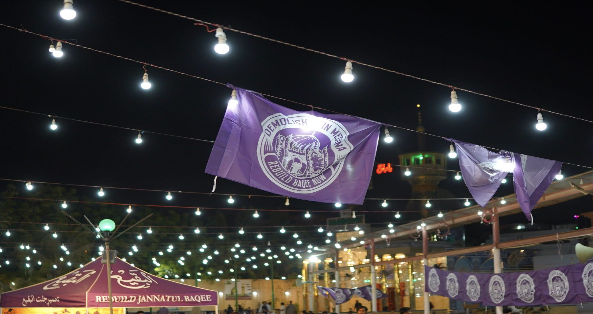 A large banner fluttering in the soft breeze reading Rebuild Baqee Now with strings of lights