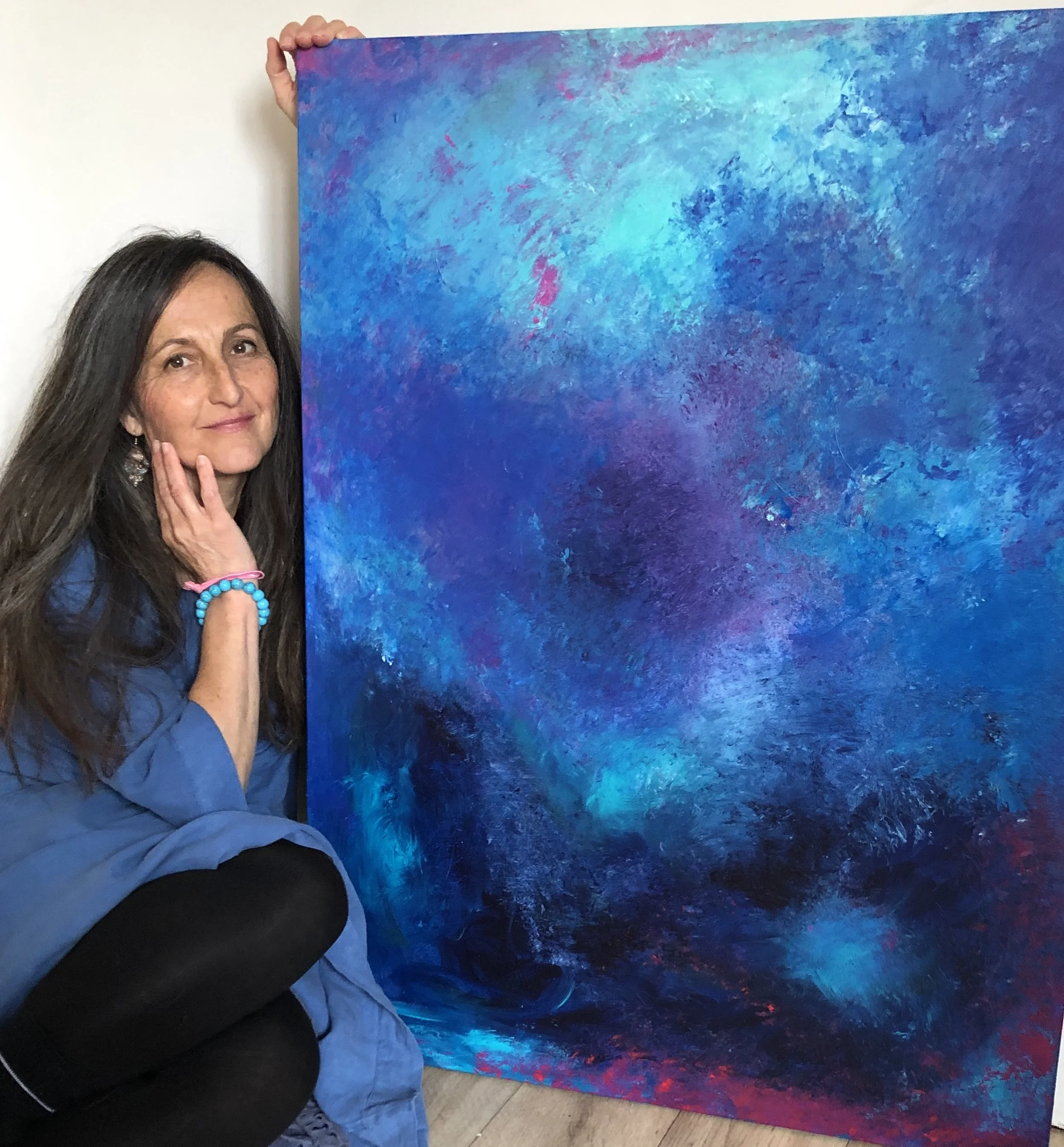 A woman with long dark hair, wearing a blue shirt, black leggings, and blue and pink bracelets, kneeling next to an abstract blue and purple painting.