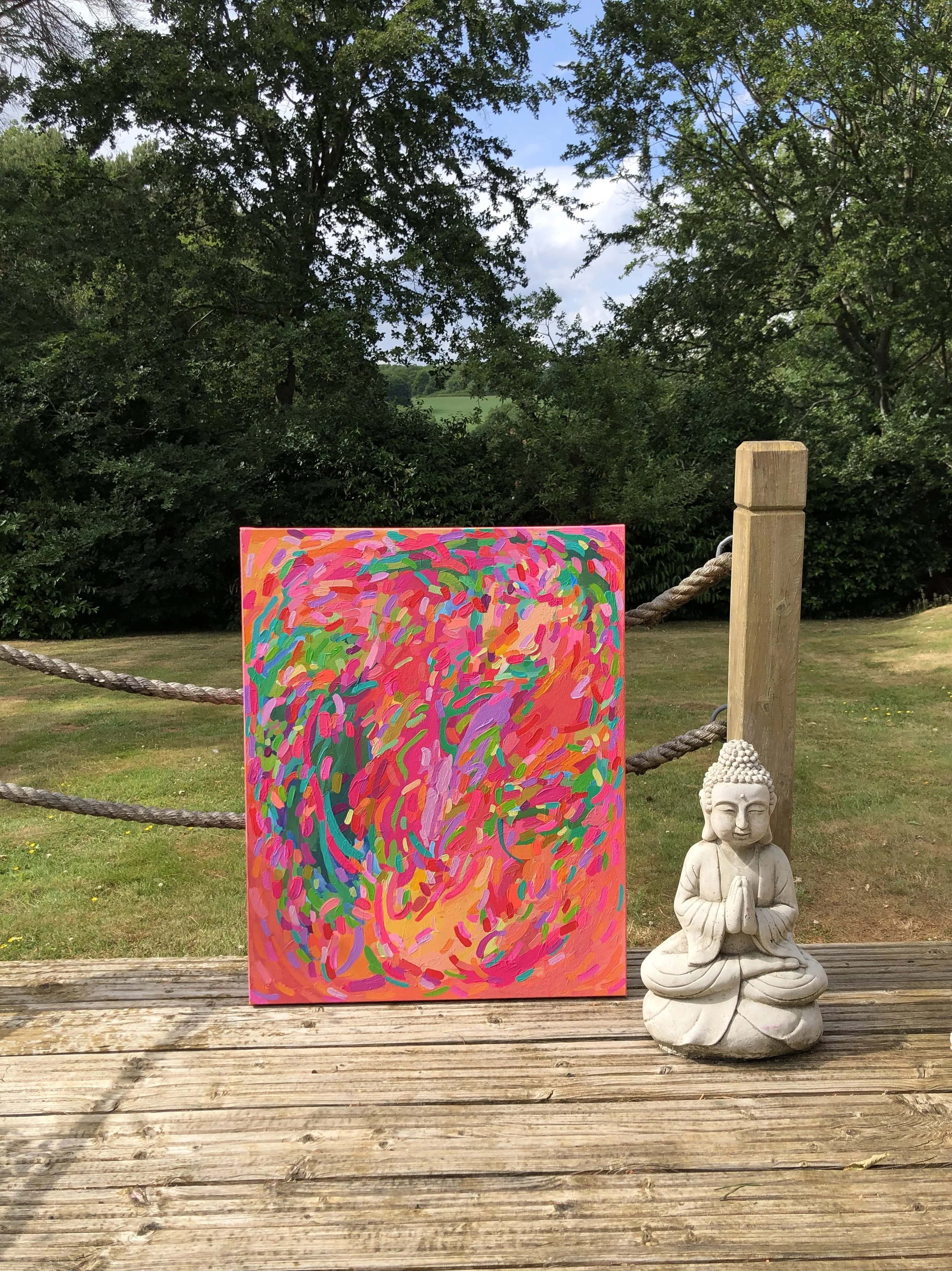 A colorful abstract painting with swirls of pink, purple, green, and orange, placed on a wooden deck outside. Next to the painting, there is a small white Buddha statue sitting in a meditative pose with hands together. Behind them, there is a wooden 