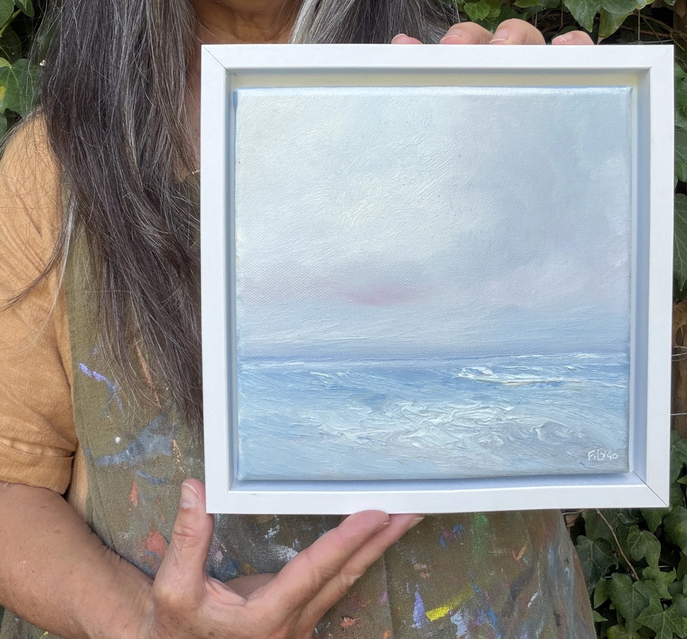 Person holding a small framed painting of a seascape with a blue sky and ocean, outdoors surrounded by greenery.