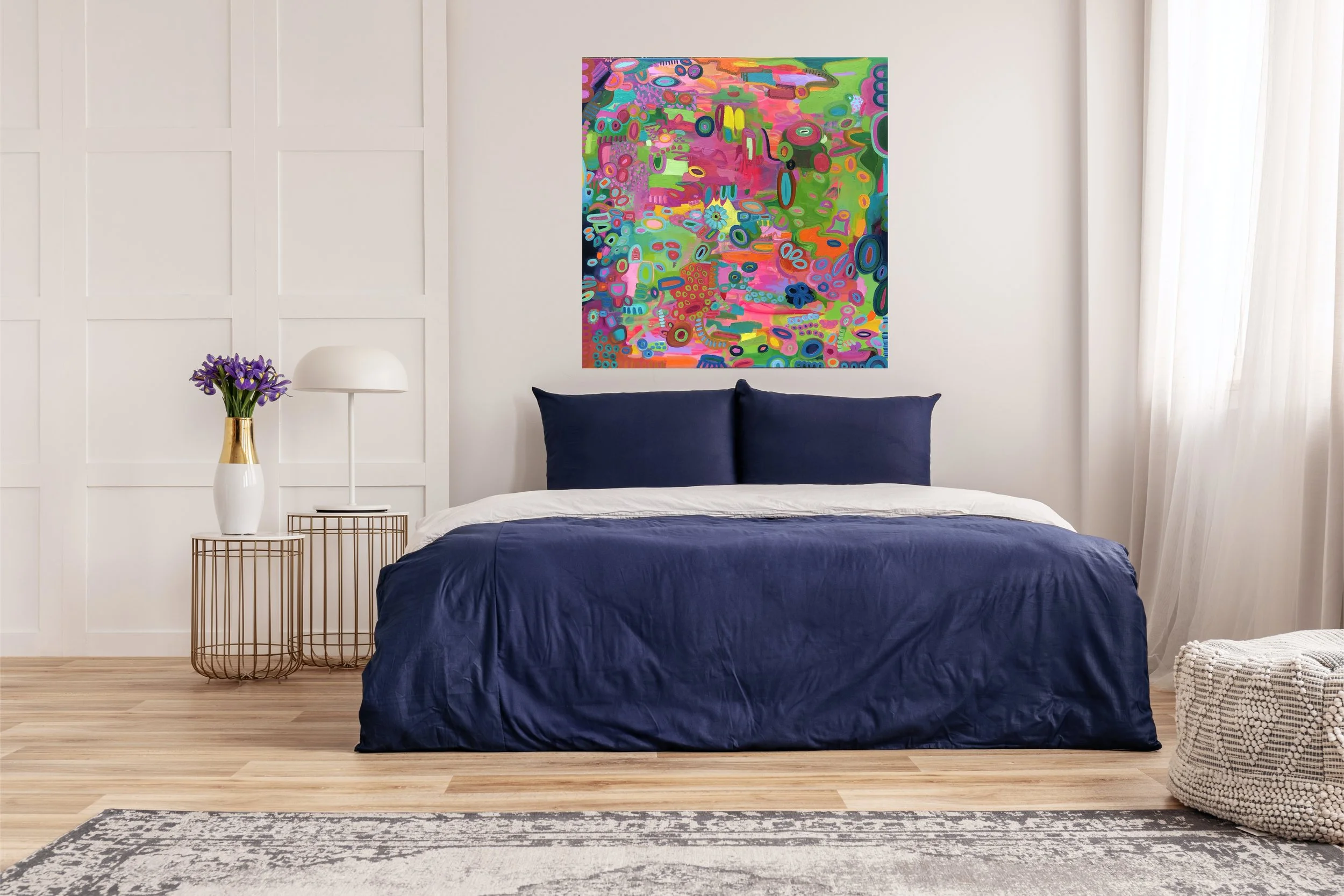 Modern bedroom with navy blue bedding, white walls, and a colorful abstract painting above the bed. There is a side table with a white vase of purple flowers and a white lamp, and a woven basket next to a window with sheer curtains.