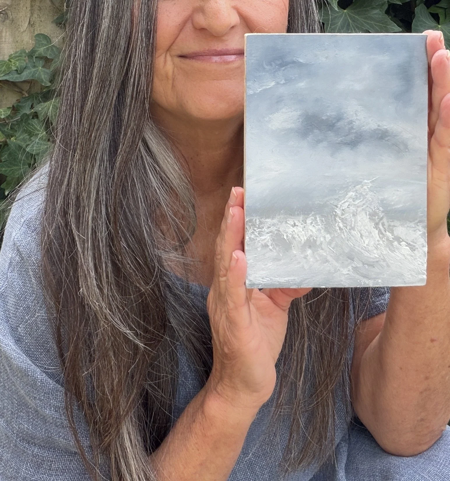 A woman with long brown hair holding a small canvas painting of a cloudy sky in front of her face outdoors, with green leaves and a wooden fence in the background.