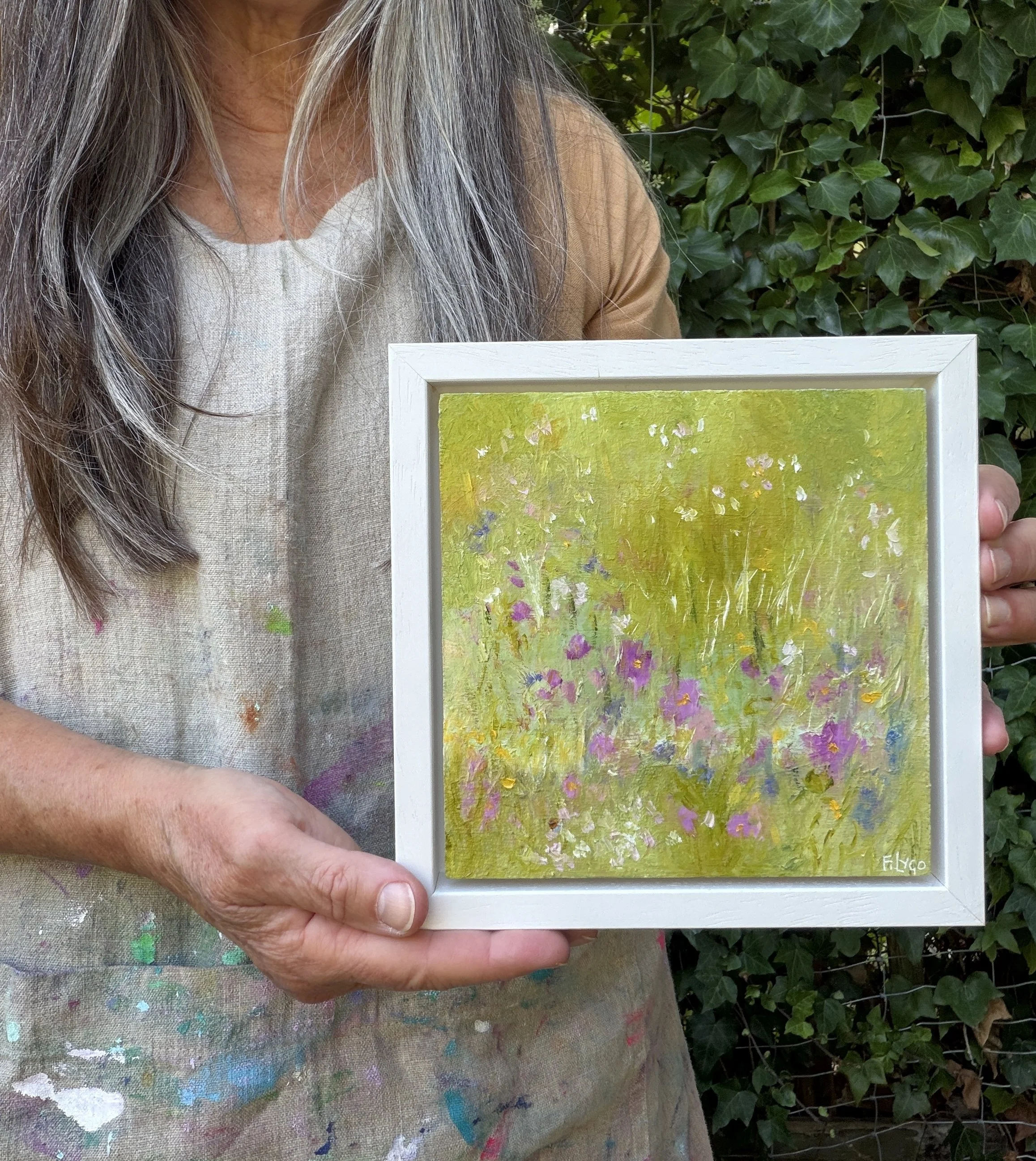 A woman with gray hair wearing a paint-splattered shirt holding a framed painting of a field with flowers against a green foliage background.