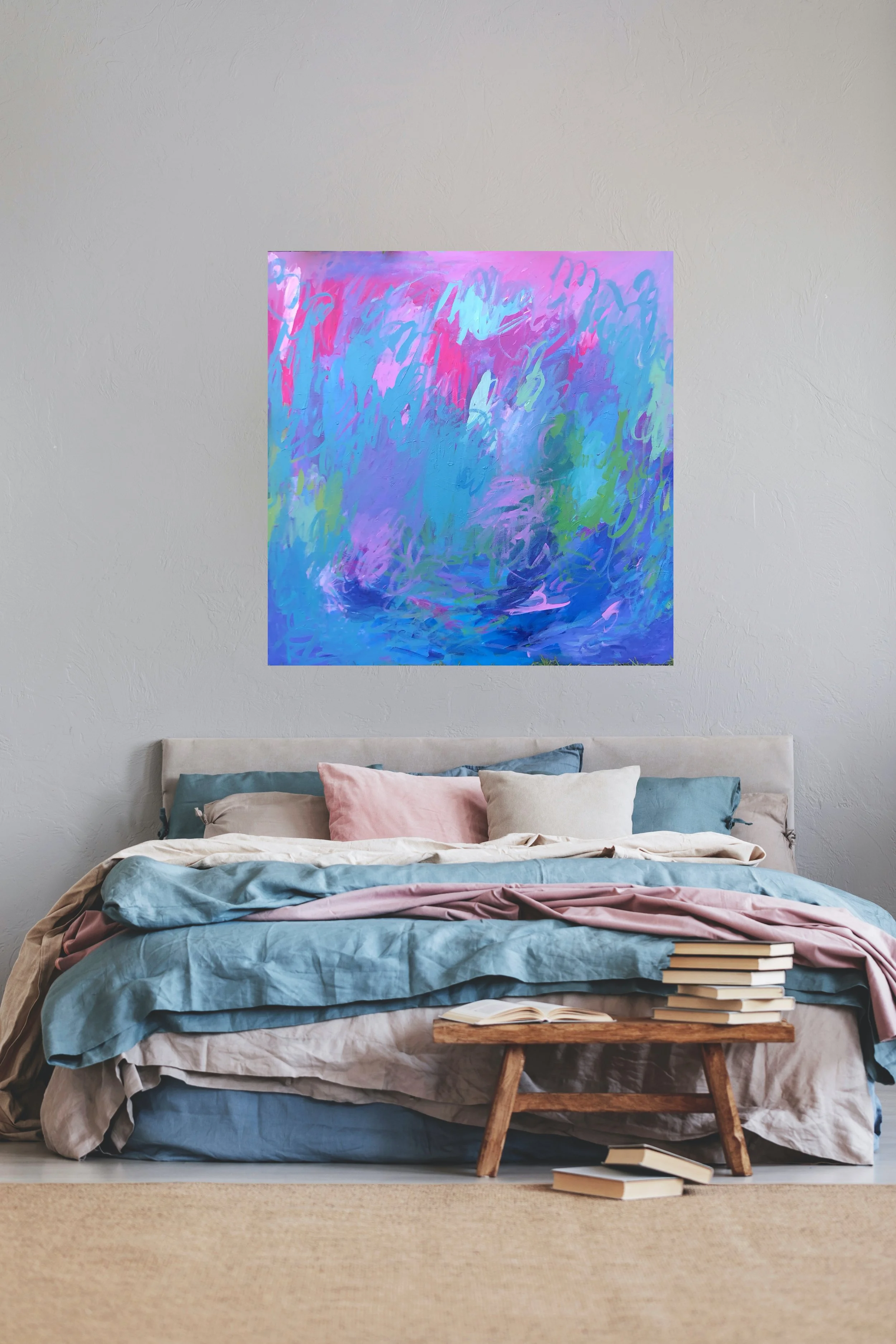 A bedroom with a beige bed and multiple pastel-colored pillows under a pink, purple, blue, green, and white abstract painting on a white wall. A small wooden table with open books and stacked books at the foot of the bed. The room has a neutral-color