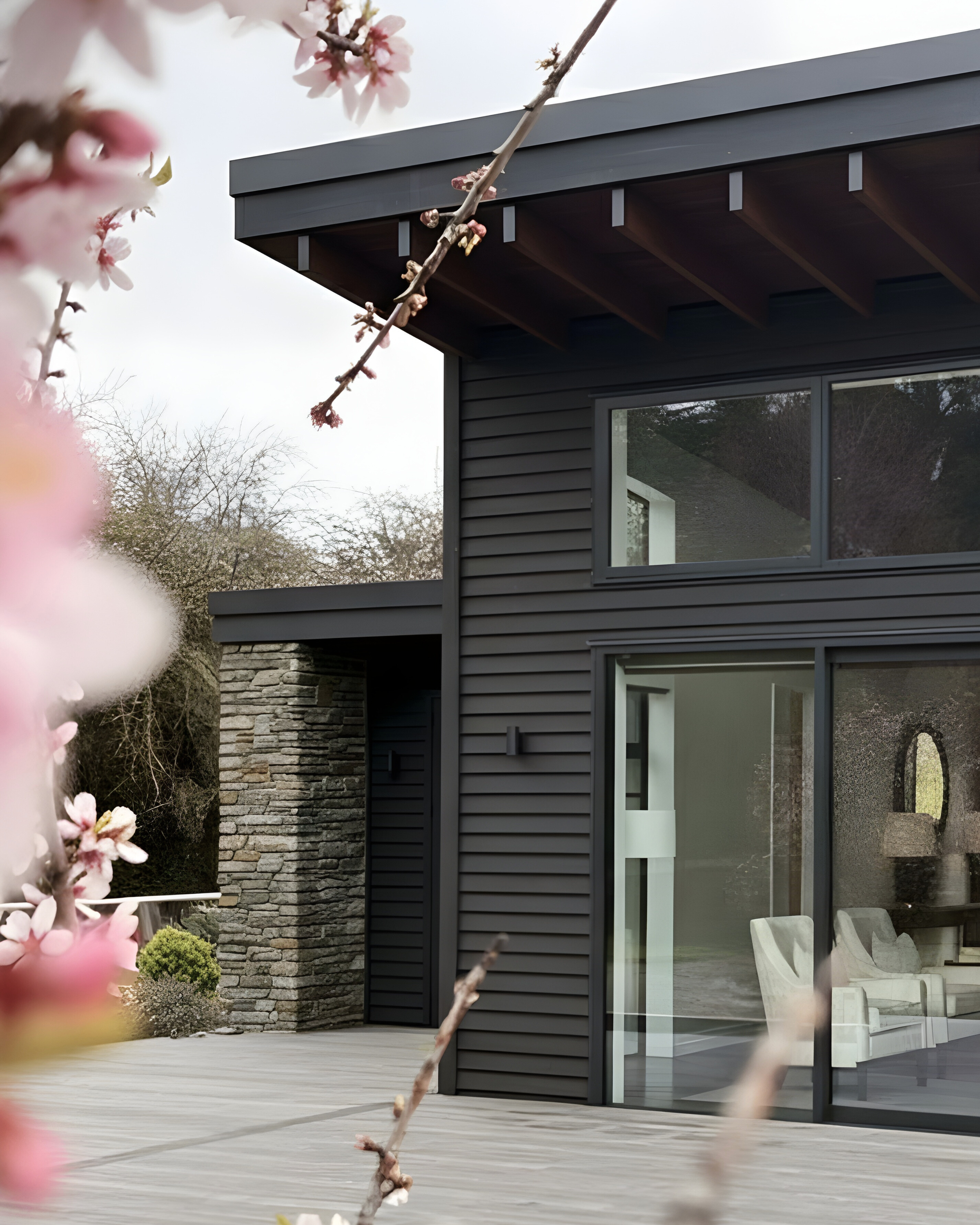 Modern black house with slatted facade, surrounded by trees and bushes, with a paved driveway and metal fence.