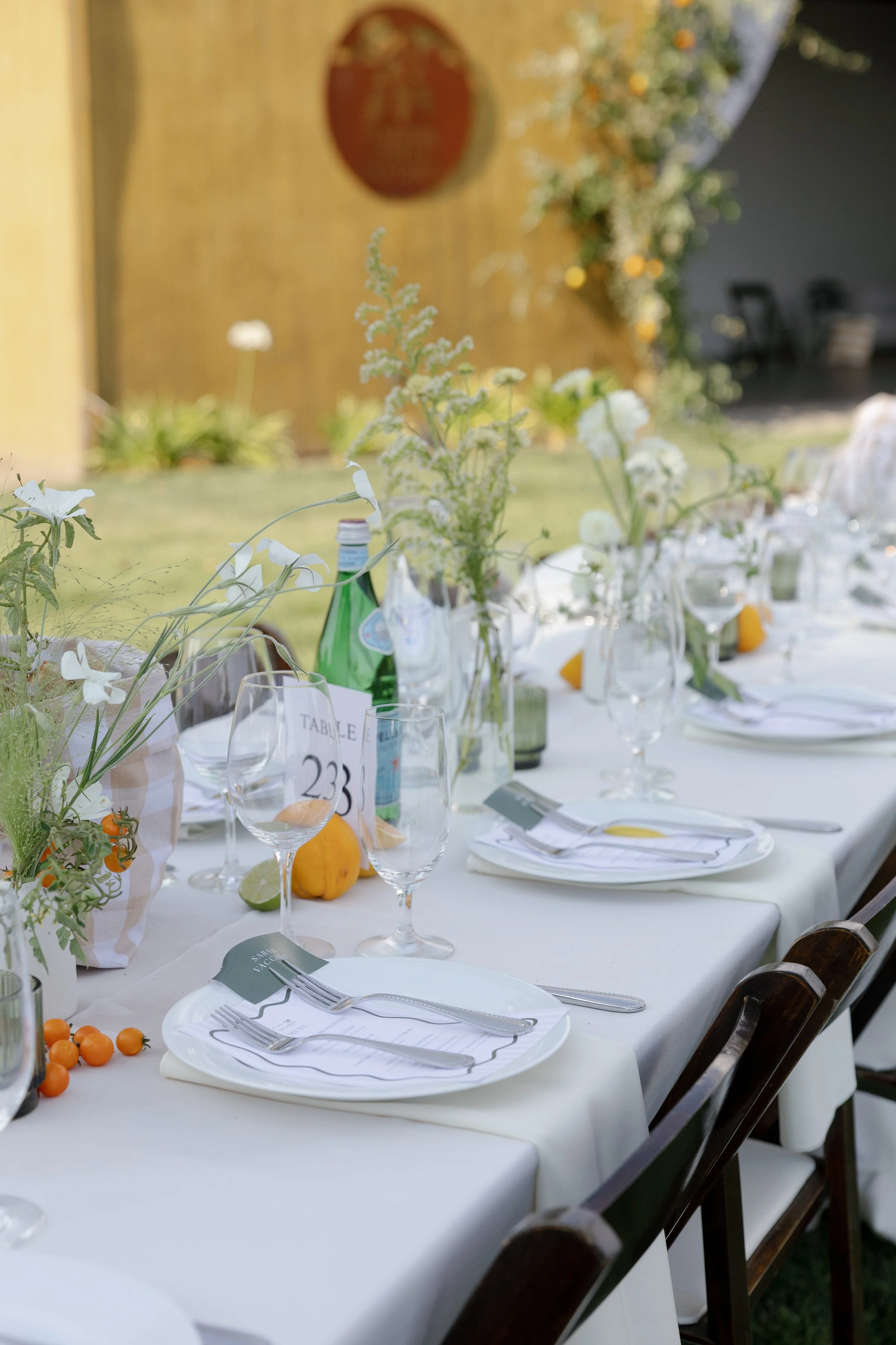 Elegant outdoor dining table set with white tablecloth, decorative floral centerpieces, glassware, plates, silverware, and small decorative gourds. The background features a wooden wall and green plants.