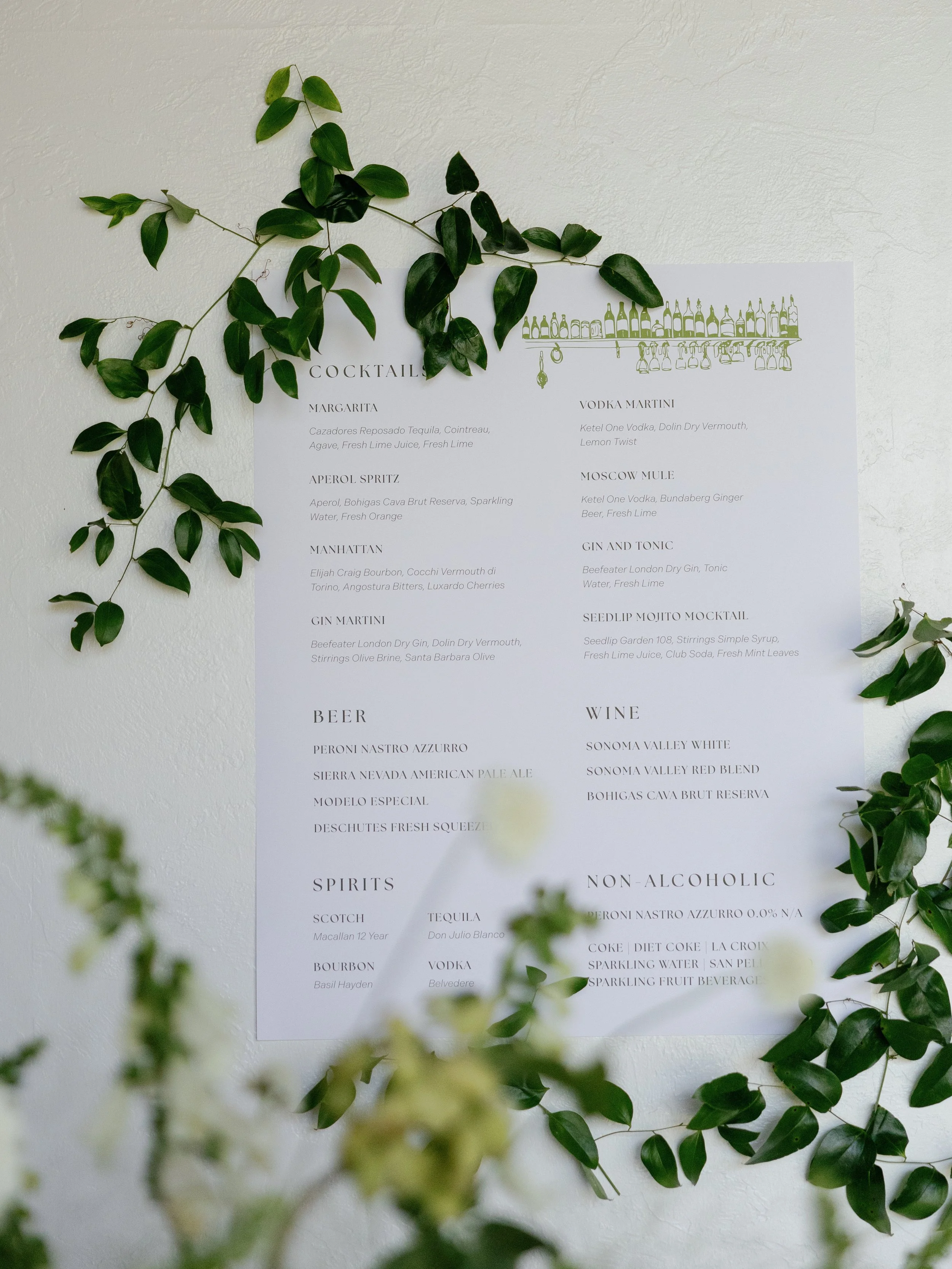 A white menu on a wall with a green vine decoration around it, listing cocktails, beer, spirits, wine, and non-alcoholic drinks.