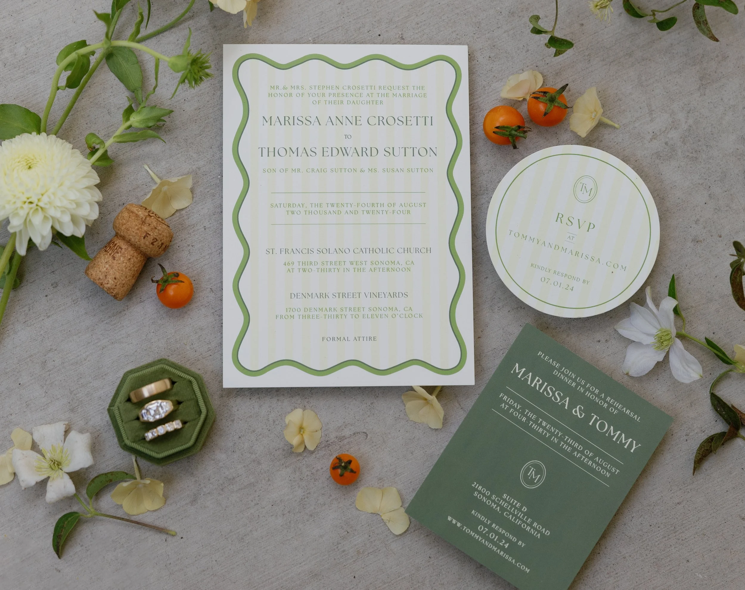 Wedding invitation with RSVP card, rings in a green ring box, small tomatoes, white flowers, and a cork, all arranged on a gray surface.