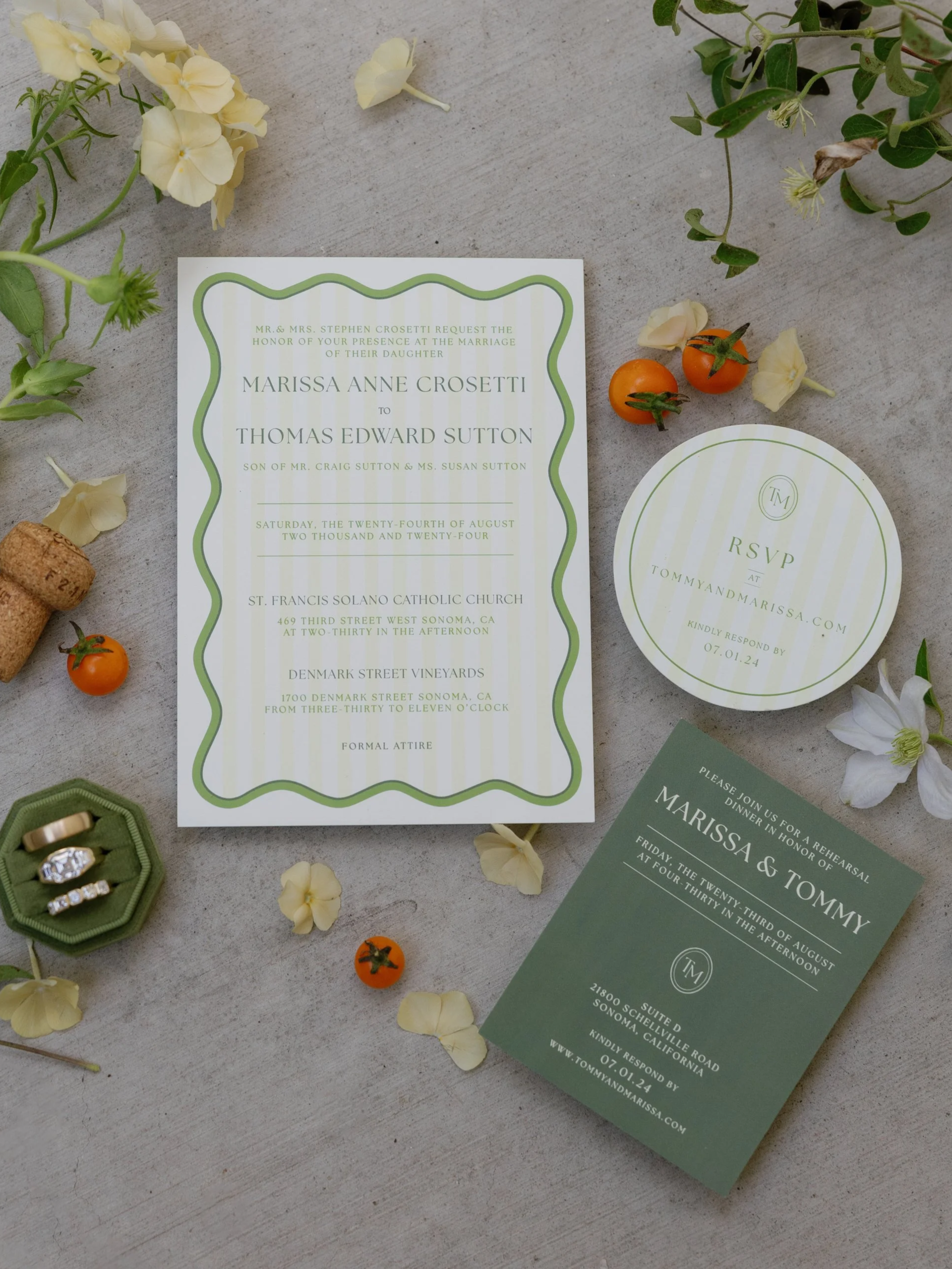 Wedding invitation suite with floral accents. The main invitation has a green wavy border and details for the wedding of Marissa Anne Crosetti and Thomas Edward Sutton, scheduled for August 24th, 2024, at two California locations. Additional RSVP and rehearsal dinner cards feature matching green and white striped and solid backgrounds. The suite is surrounded by flowers, small orange tomatoes, and a ring box with rings.