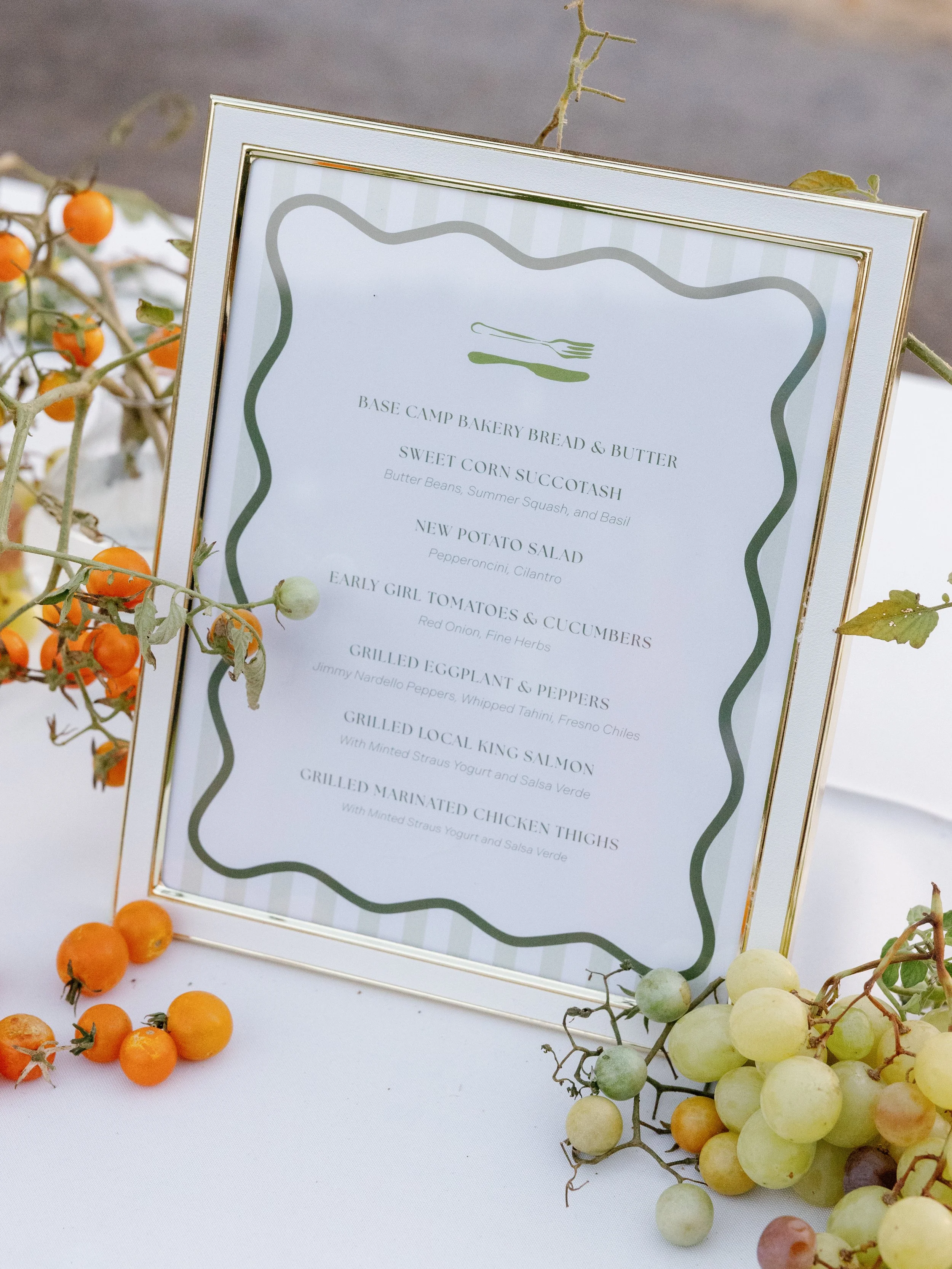Restaurant menu in a gold frame on a white table with orange and green grapes around it.