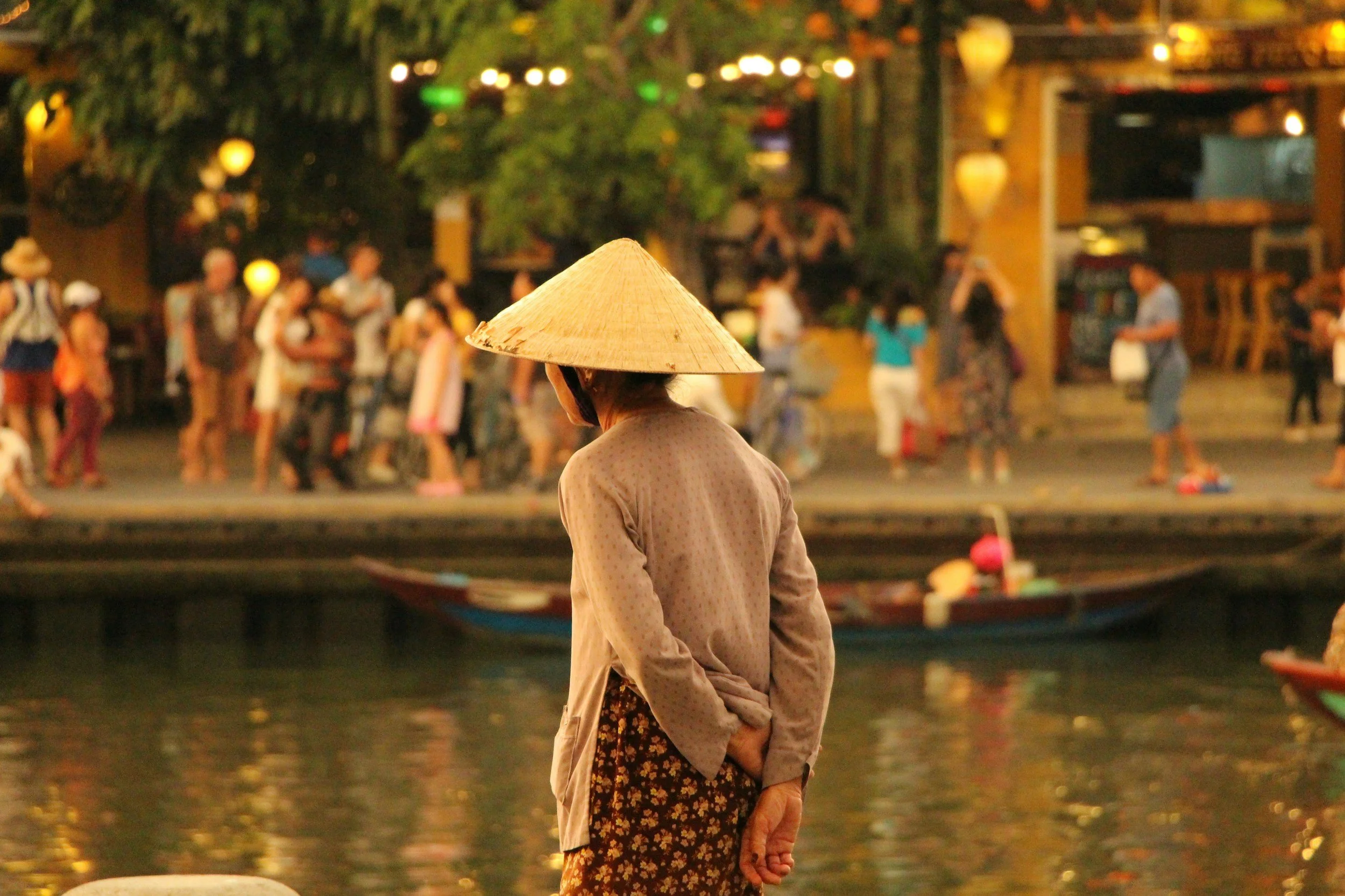 A person with a traditional conical hat standing near a body of water with people dancing and socializing in the background during the evening.