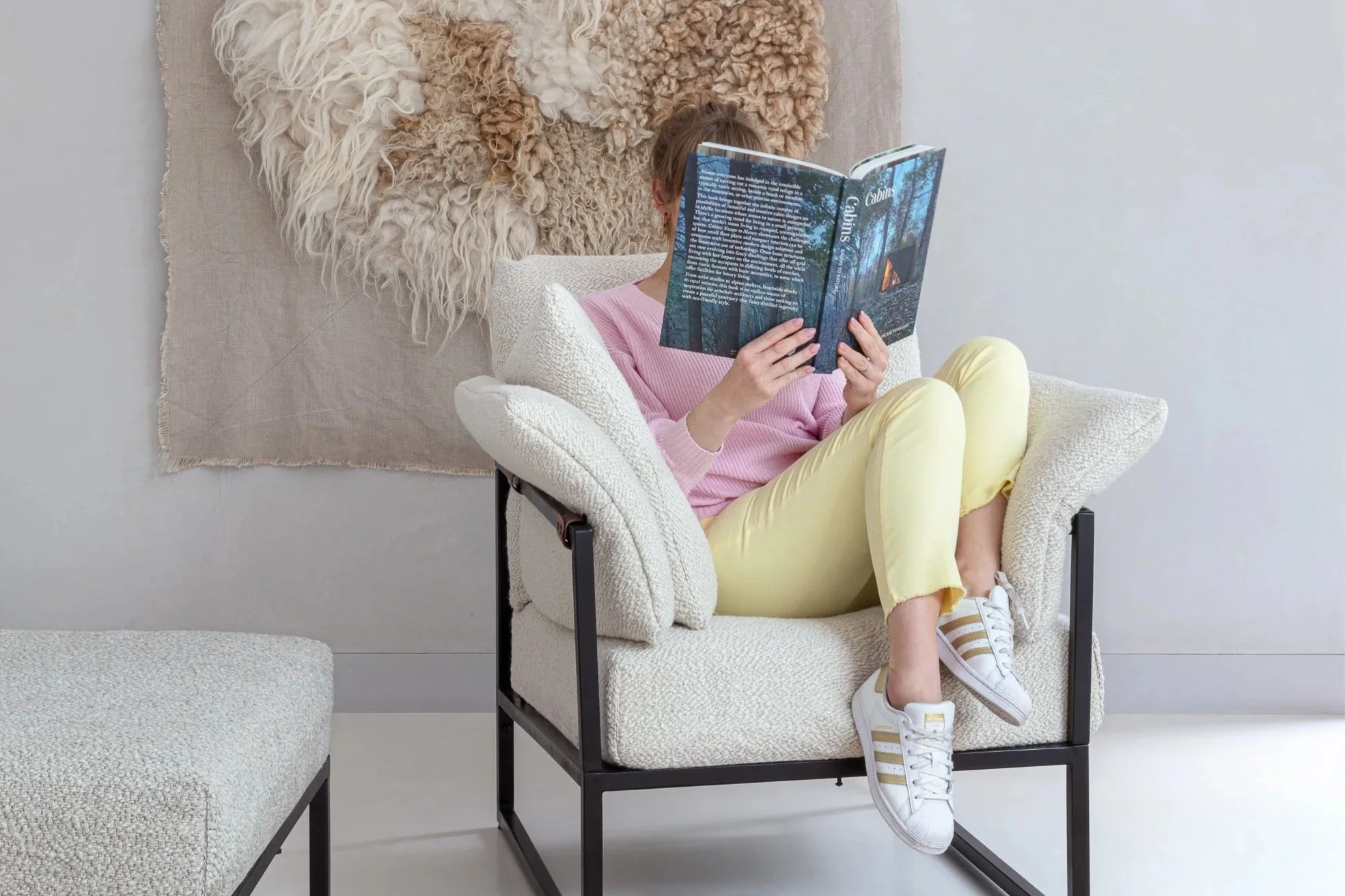 Ligne Pure Belgian multi-me sofa and rug - A person sitting on a beige armchair with black metal frame, reading a magazine. They are wearing a pink top, yellow pants, and white Adidas sneakers with gold stripes.