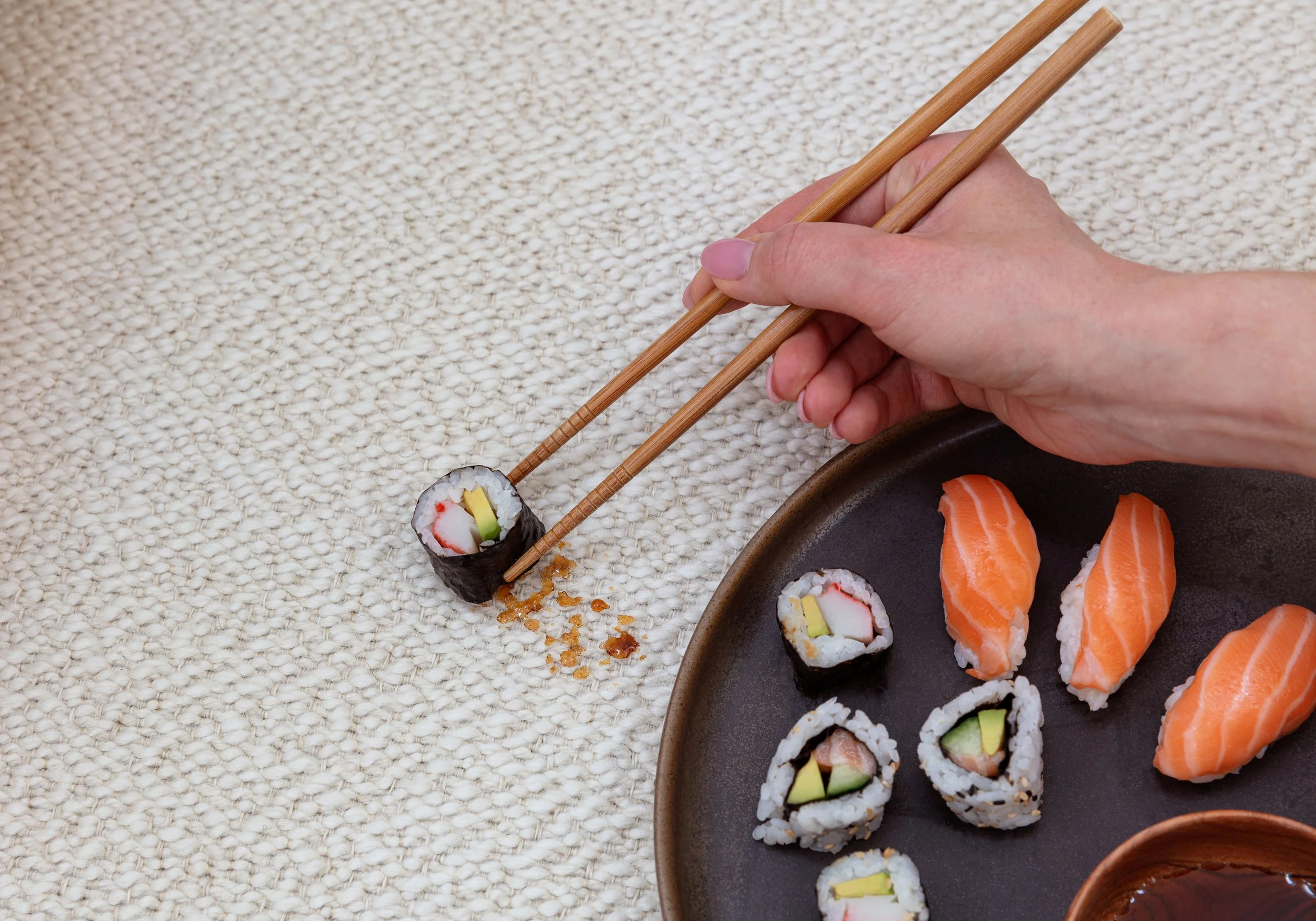 Ligne Pure Belgian multi-me sofa and rug - Person using chopsticks to pick up a sushi roll with crab stick and cucumber from a textured table, with a plate of assorted sushi including salmon and avocado rolls nearby.