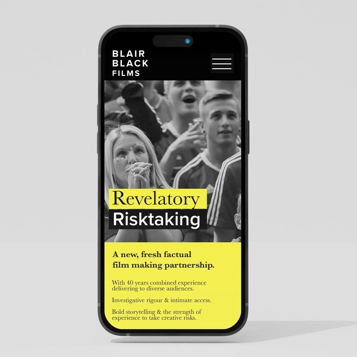 A smartphone displaying a webpage for Blair Black Films with a black-and-white photo of a group of people, mainly young adults, looking intrigued or emotional. The webpage highlights a film titled 'Revelatory Risktaking' with a brief description in yellow, mentioning a new factual film in partnership.