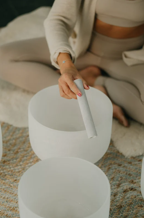 Person sitting on a rug playing a white crystal sound bowl with a mallet meant for healing and re-alignment