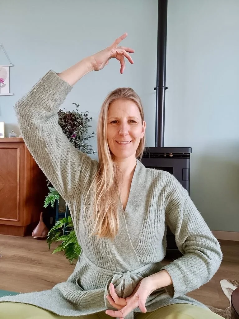 Kirsten Jense in een groene trui en broek, zittend in een meditatieve houding met een hand boven haar hoofd en de andere in een mudra. Energie lichaamswerk en bewustzijnsontwikkeling in een rustgevende setting.