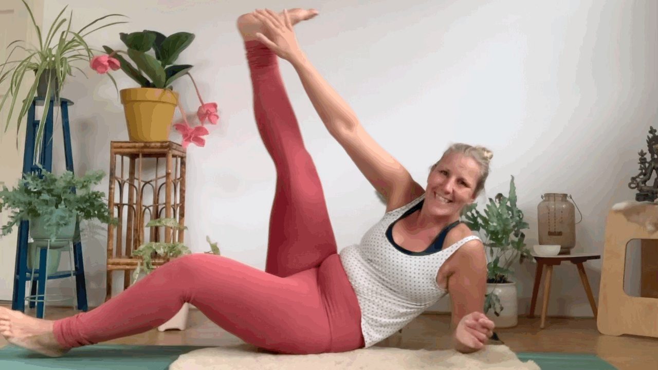 Kirsten Jense lachend op een yogamat in een yogahouding, met één been omhoog. Ze draagt een roze broek en witte top, haar blonde haar in een knot. Witte achtergrond met groene planten en roze bloemen.