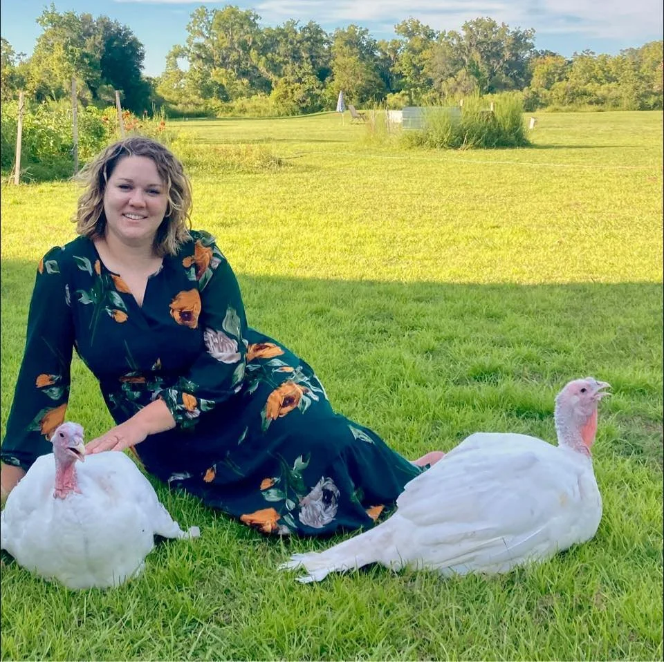 A woman sitting on a grassy field with two white turkeys, one on each side of her. She is smiling and wearing a dark floral dress. The background shows trees and a clear sky.