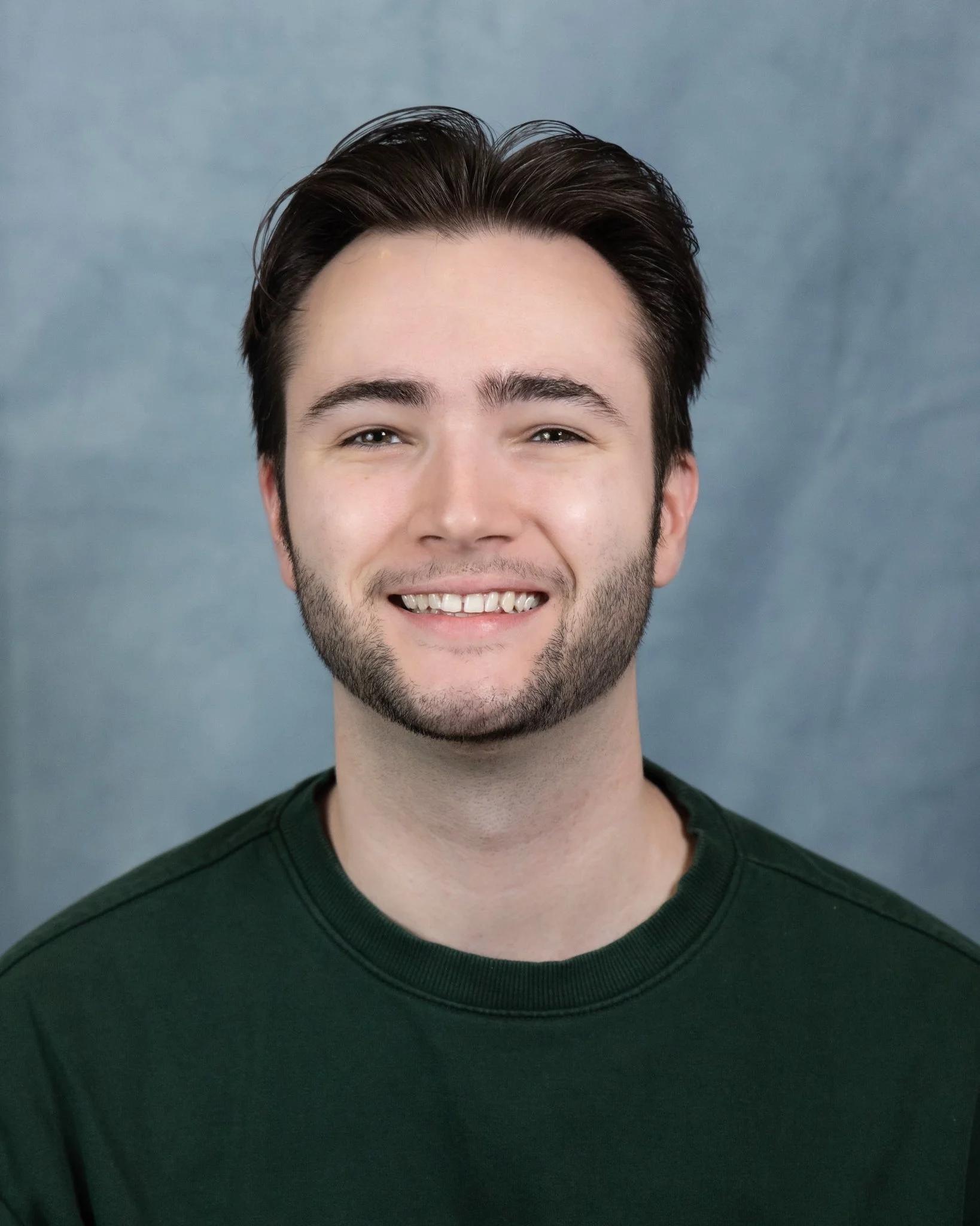Close-up photo of a young man smiling, showing his teeth, with dark brown hair parted and styled back, wearing a dark green crew neck shirt, against a plain gray background.