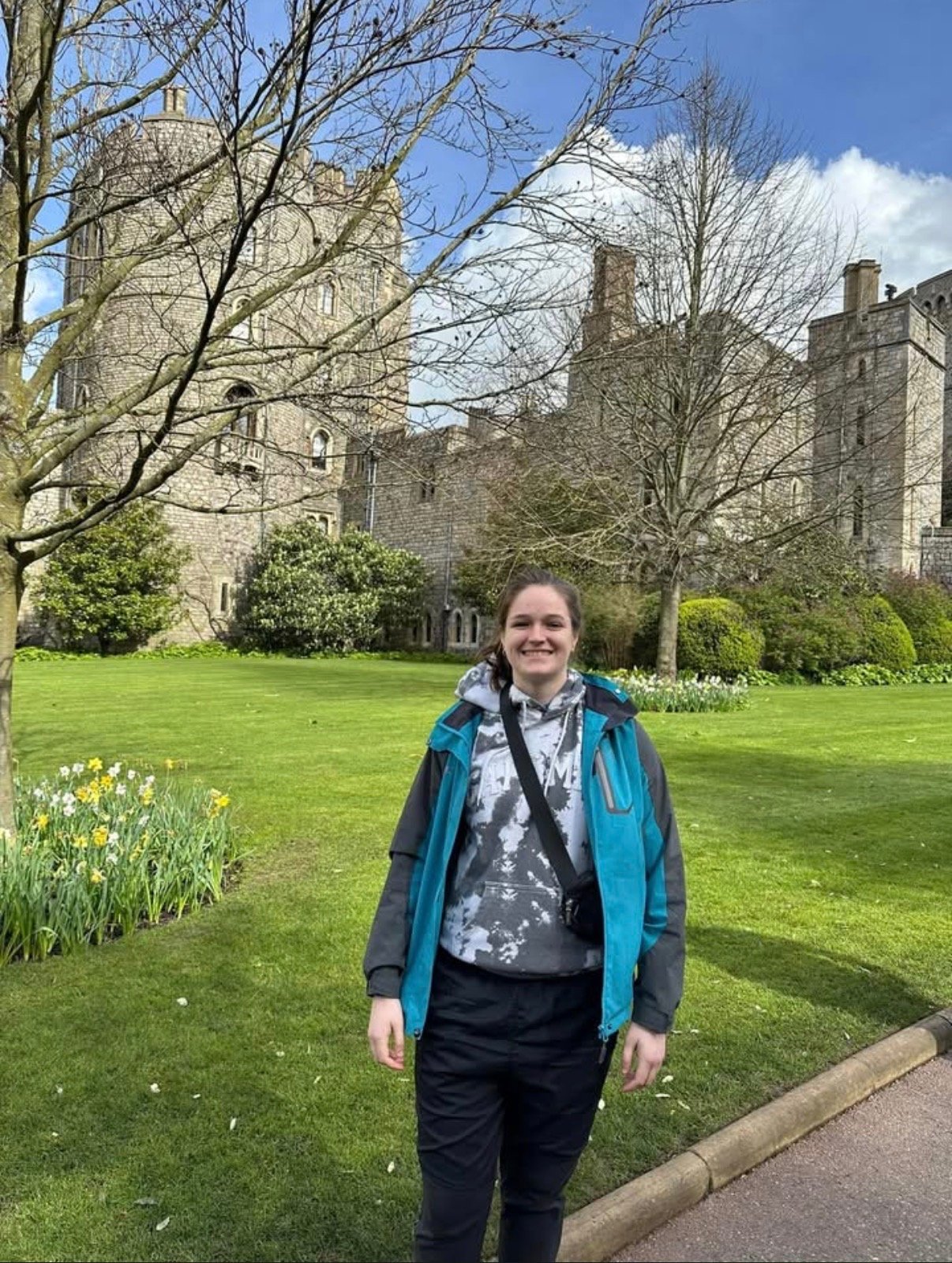 A smiling young woman in a blue and black jacket standing on a neatly trimmed grass lawn with blooming daffodils, leafless trees, and a castle-like stone building in the background under a partly cloudy sky.