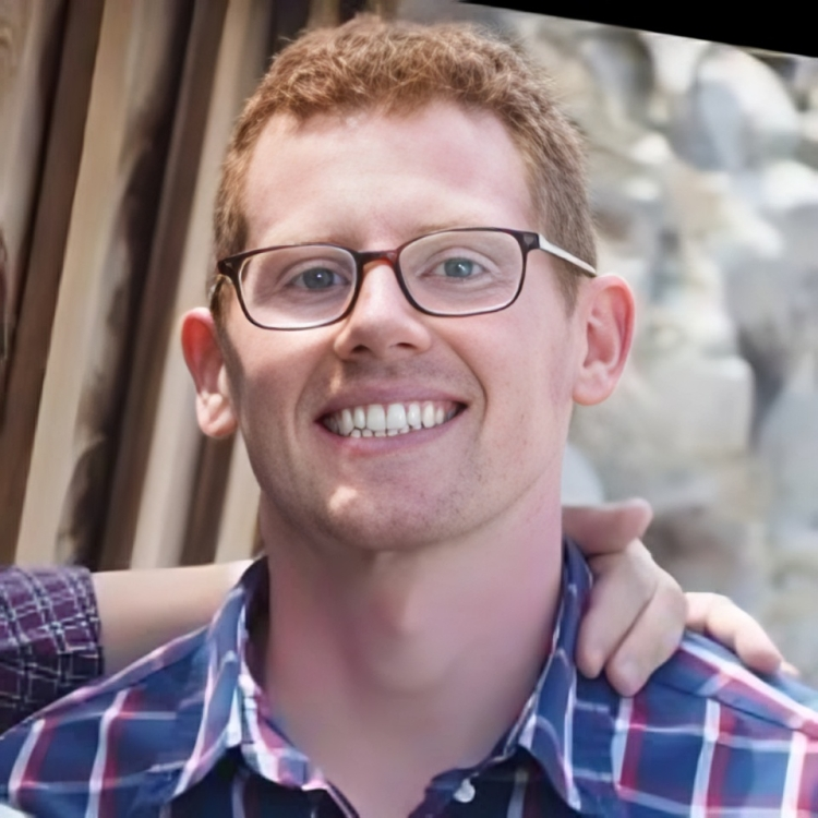 A smiling man with glasses and short red hair, wearing a plaid shirt, standing outdoors, with a hand on his shoulder.