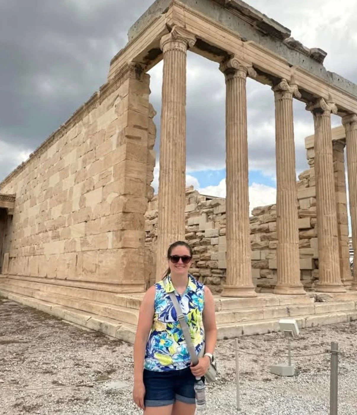 A woman wearing sunglasses, a colorful sleeveless shirt, and shorts, standing in front of ancient Greek ruins with tall columns and stone walls, under a cloudy sky.