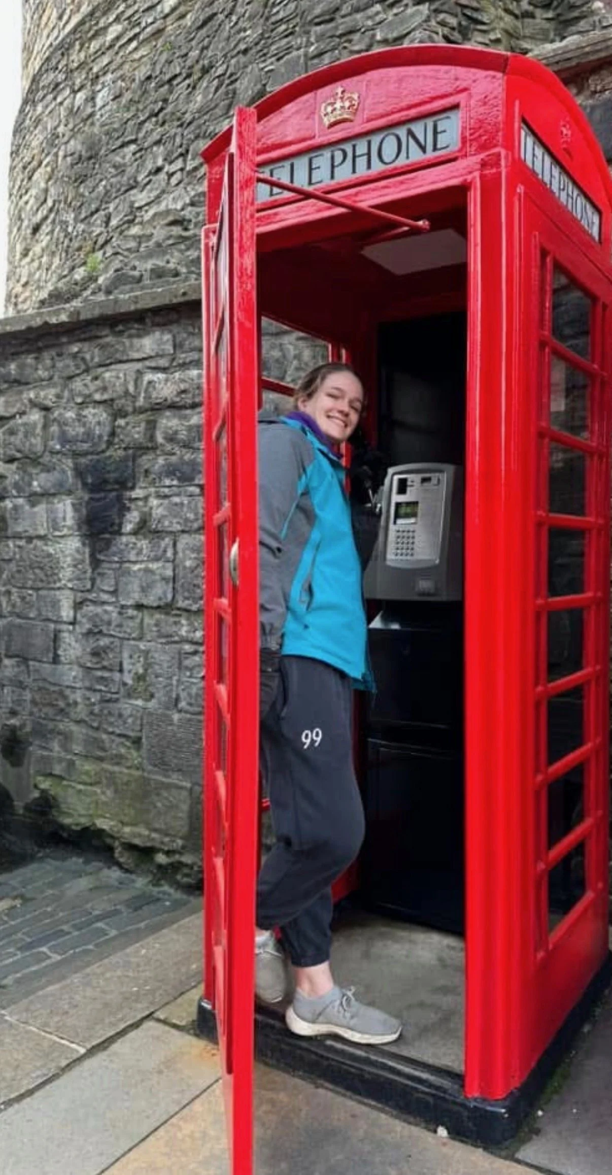 A woman smiling inside a red British telephone booth, using a public payphone on a stone street.