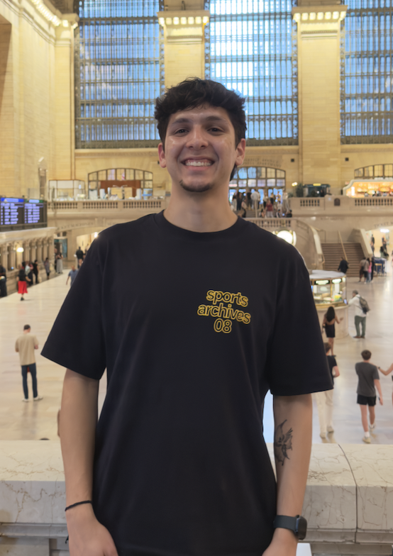 Young man smiling inside a grand, historic train station with large windows and busy travelers in the background.