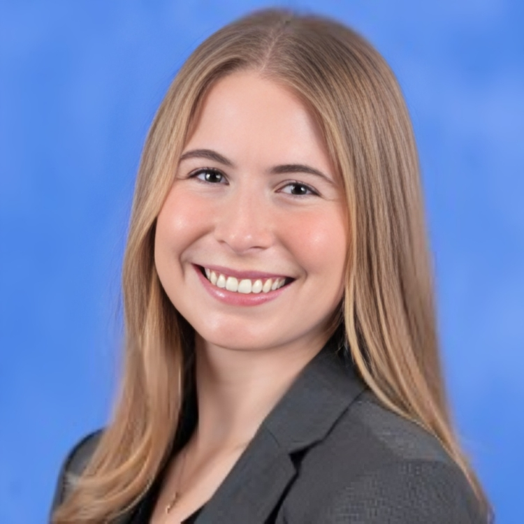 A young woman with long blonde hair smiling in front of a blue background, wearing a dark blazer.