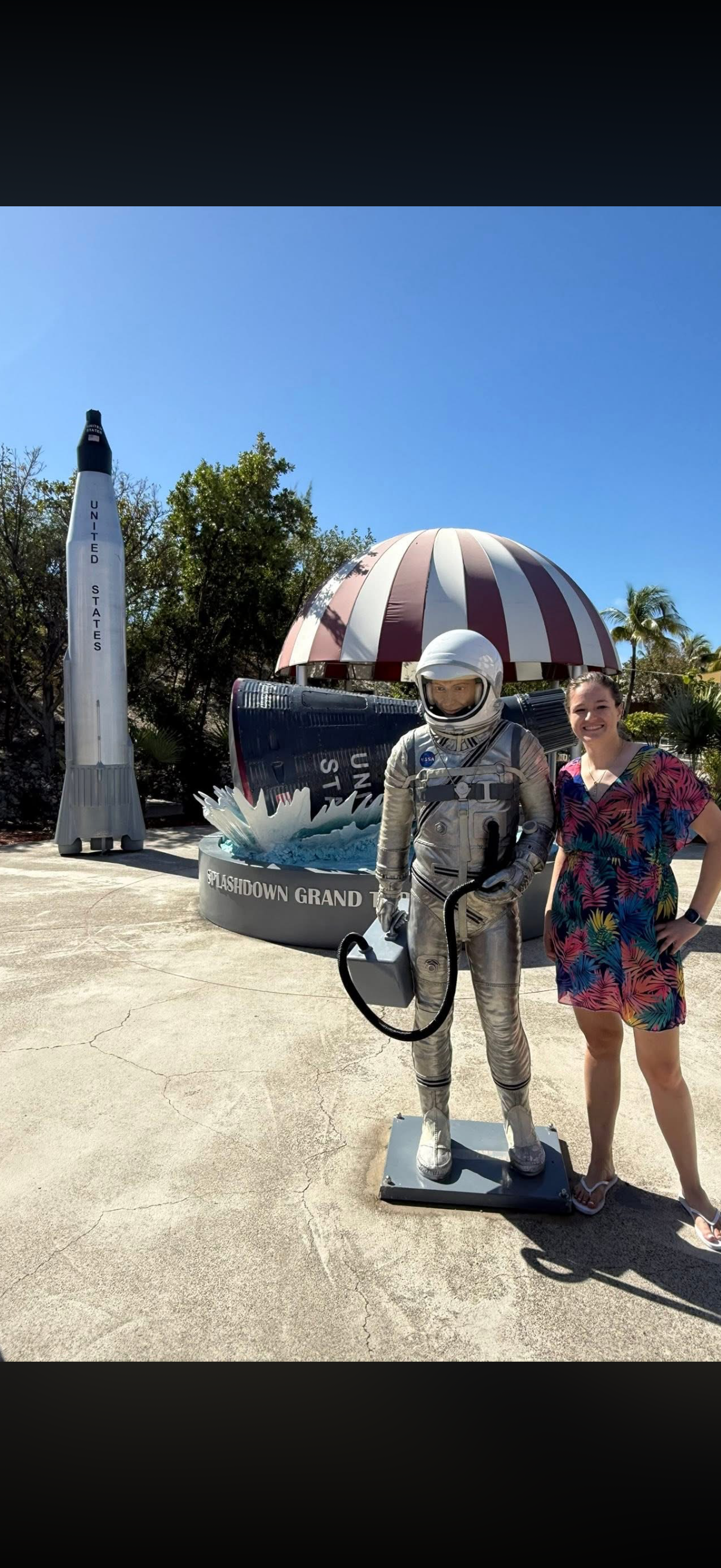 Woman in colorful dress posing next to astronaut statue holding a space helmet at Space Lashdown Grand opening with rocket and space-themed decorations in the background.