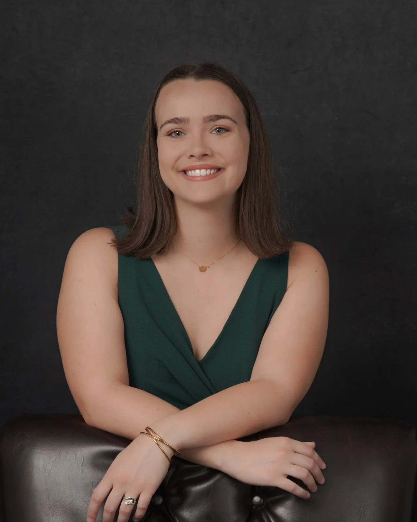Young woman with shoulder-length brown hair smiling, wearing a green sleeveless top, with her arms crossed and jewelry, against a black background.