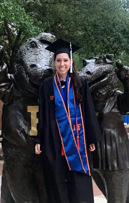 Woman in graduation cap and gown standing in front of a large bear statue at a university.